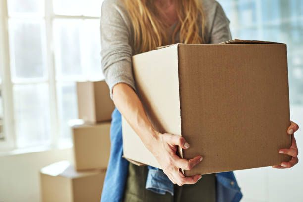 Person carrying a large moving box during a home relocation with packed boxes in bright room.