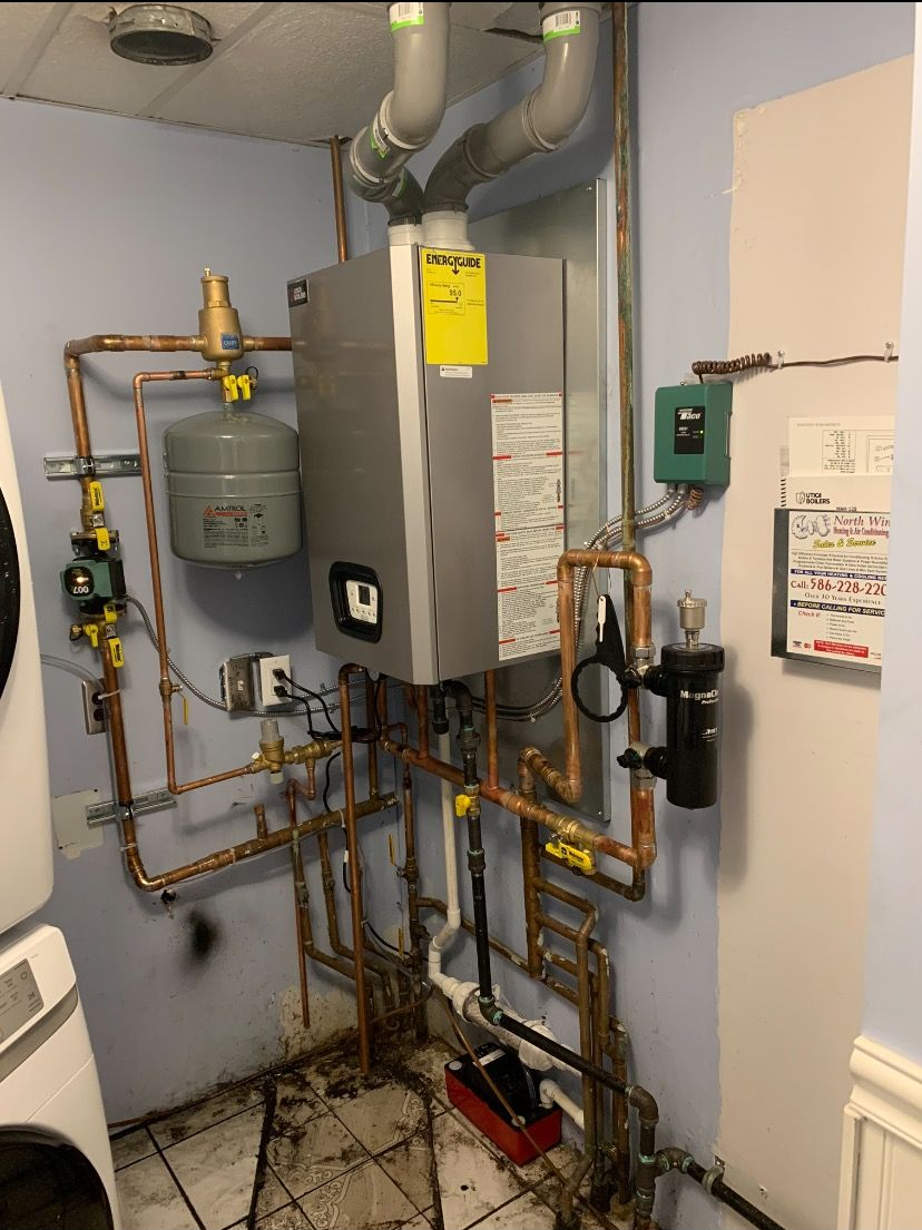 A wall-mounted combi boiler system with copper piping, a gray expansion tank, and electrical components in a utility room.