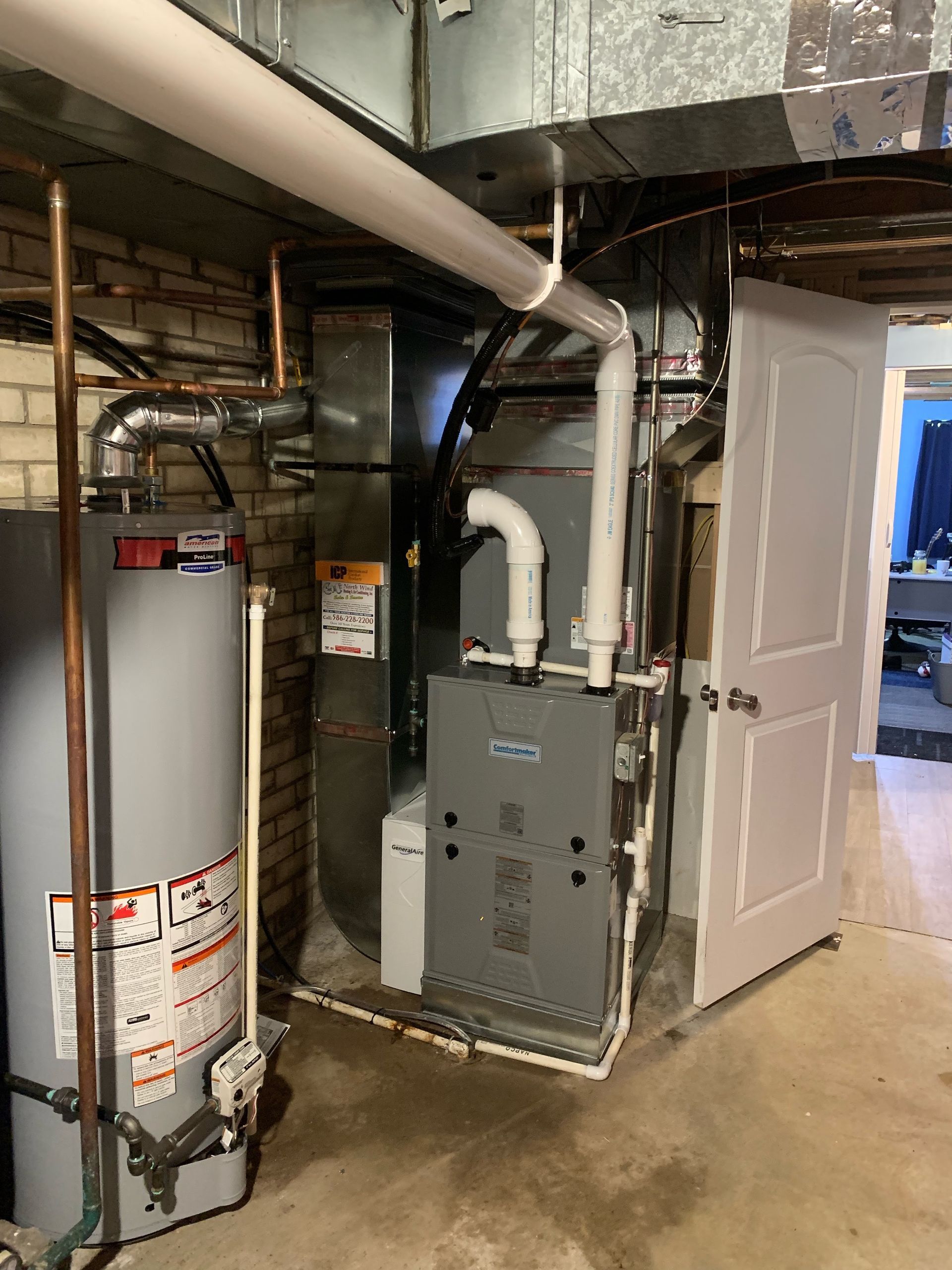 Basement with furnace, water heater, and ductwork near a white door and brick wall.