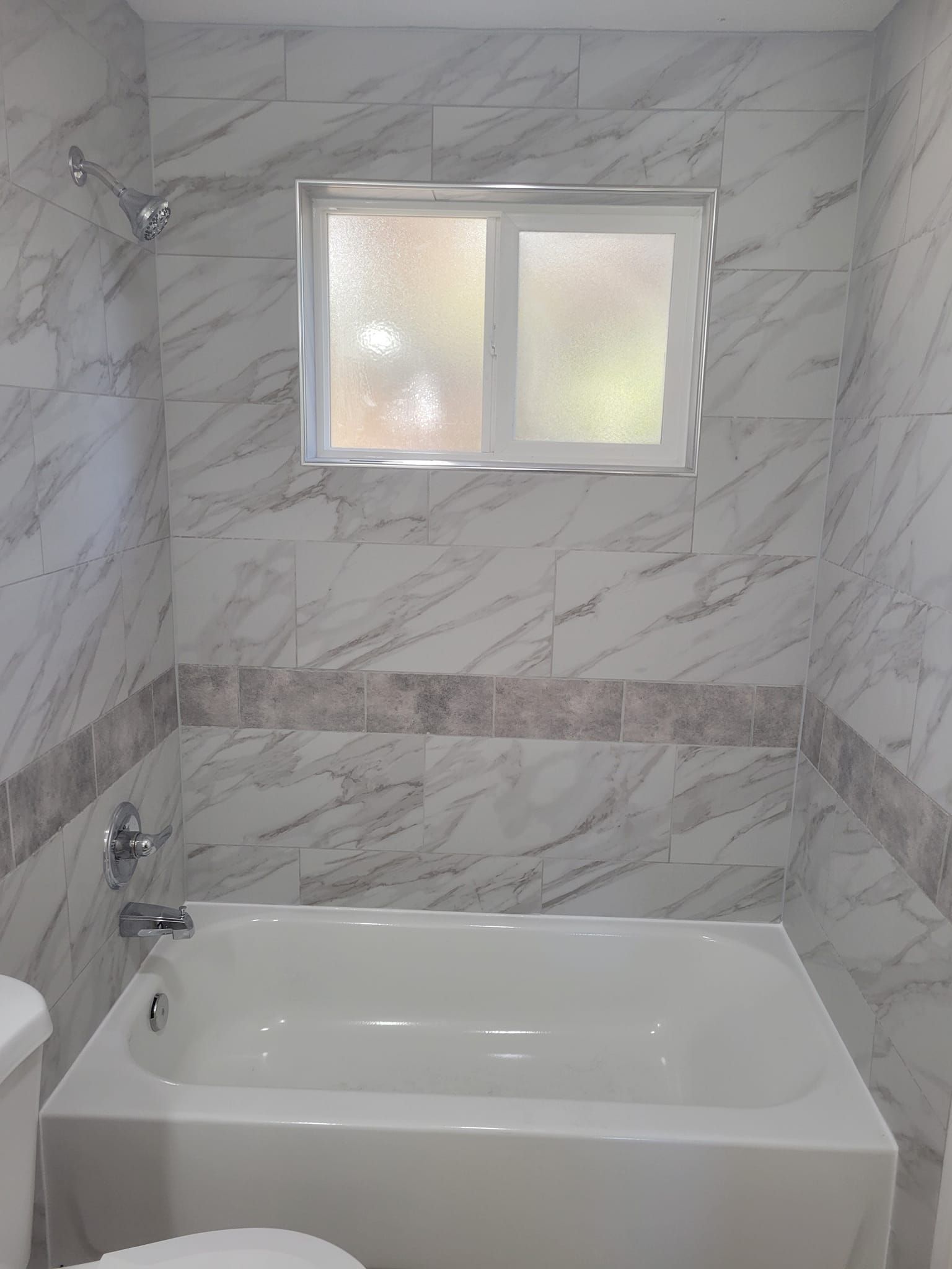 Bathroom with white tub, marble-look tiles, shower head, and frosted window.