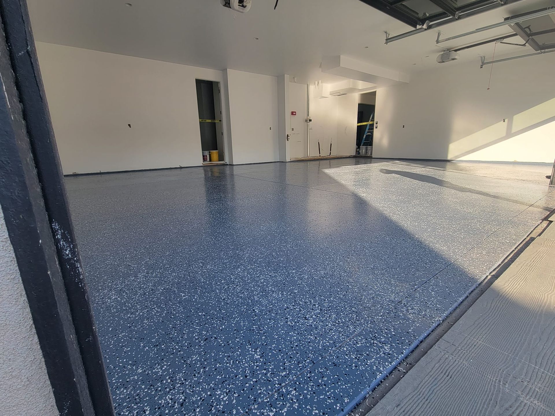 Newly coated garage floor, blue with flecks, reflecting sunlight. White walls, open door.