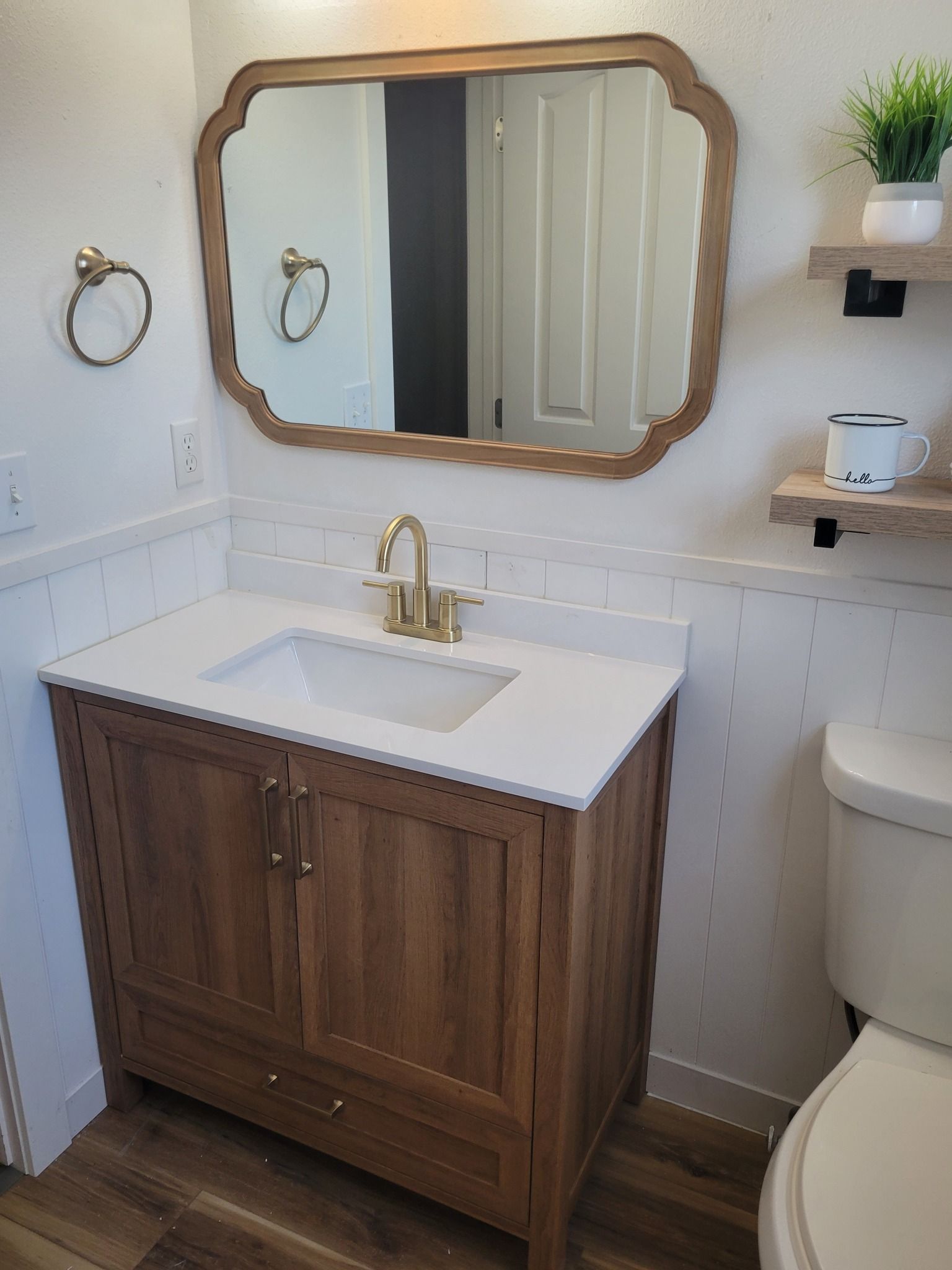 Bathroom with wooden vanity, gold fixtures, white countertop, and scalloped mirror.