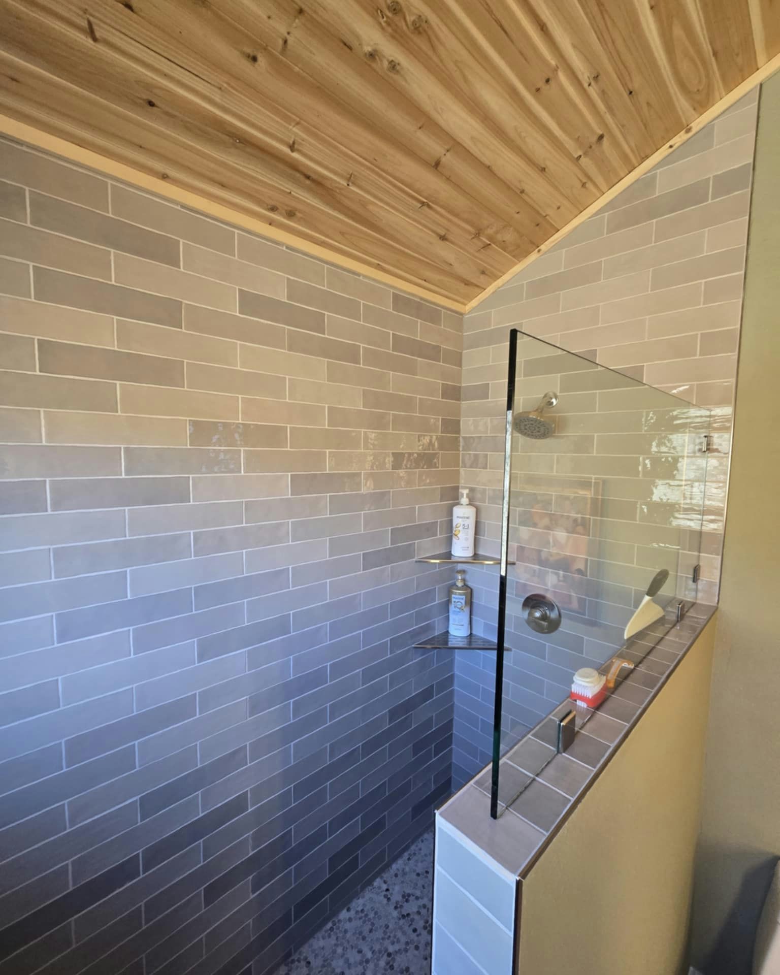 Shower with gray brick tiles, glass divider, pebble floor, and wooden ceiling.