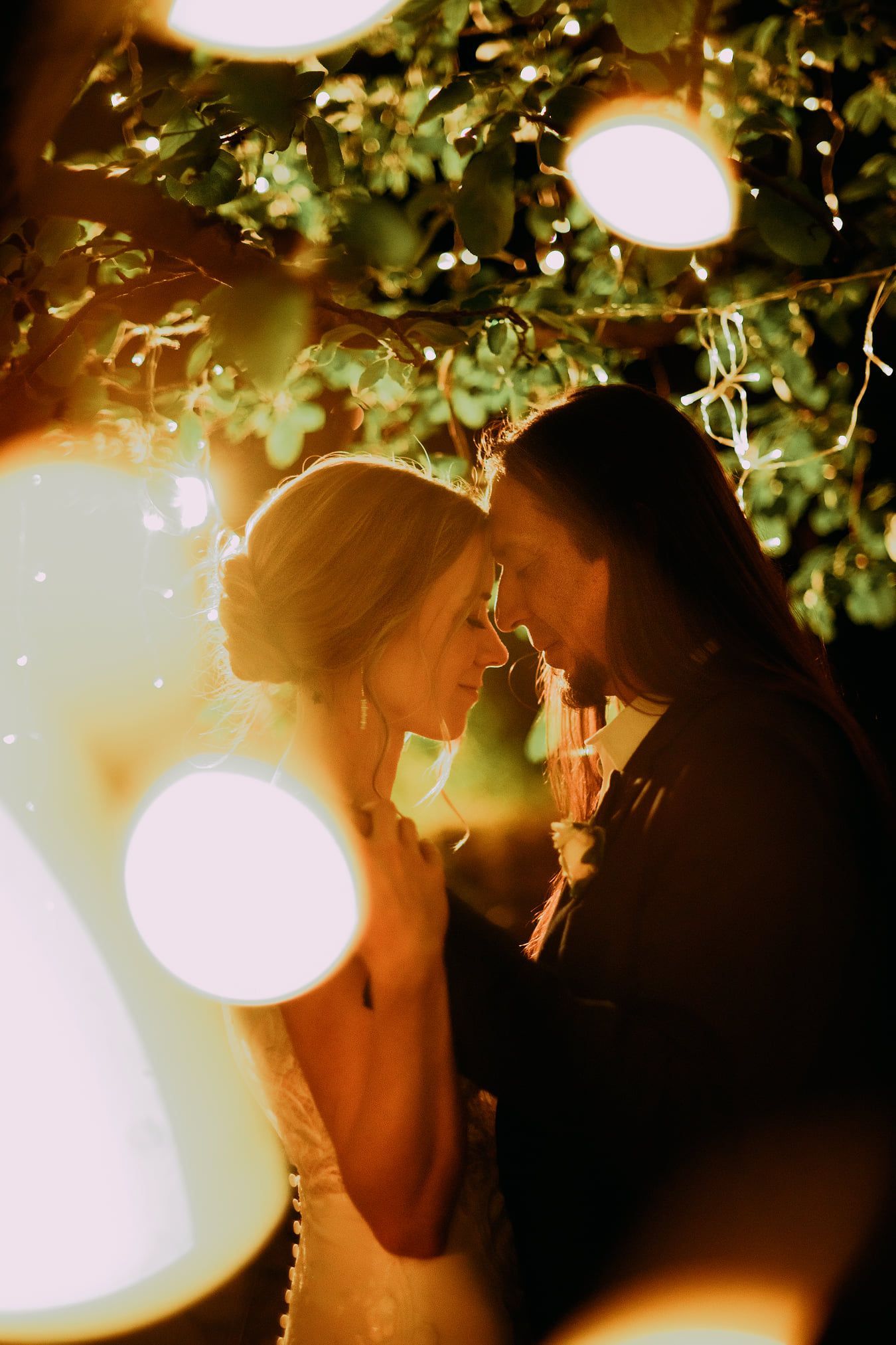 Bride and groom embrace under fairy lights in a romantic outdoor setting. Soft lighting and affectionate expressions.