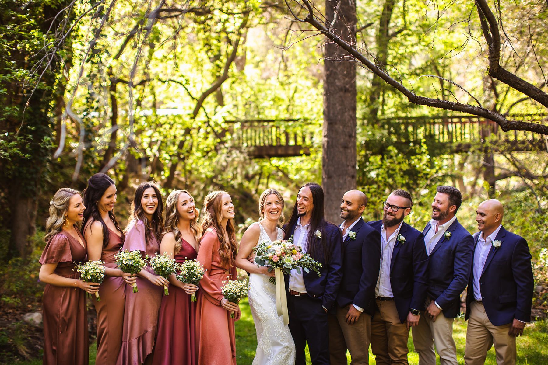 Wedding party: bride laughing with bridesmaids in rose dresses, groomsmen in suits, outdoor setting.