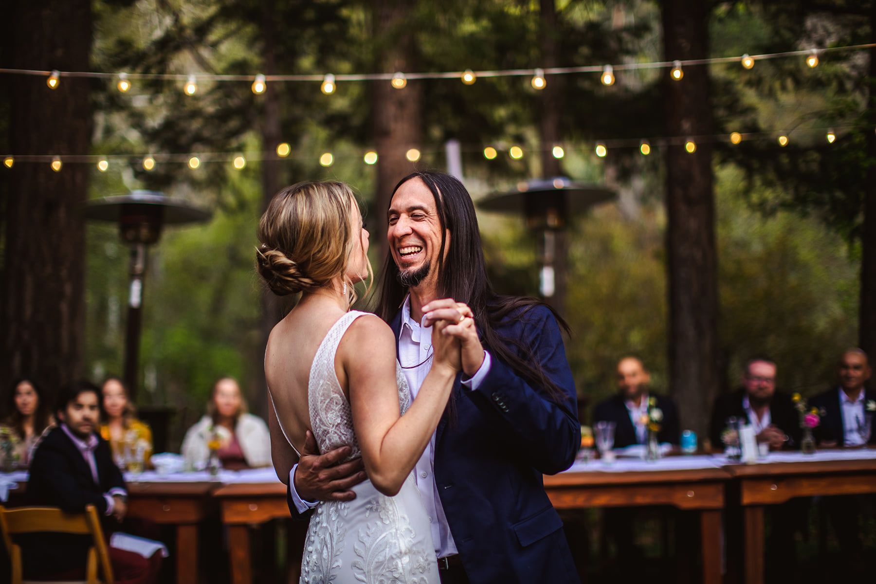 Bride and groom dance outdoors at their reception. They smile at each other, surrounded by guests and strung lights.