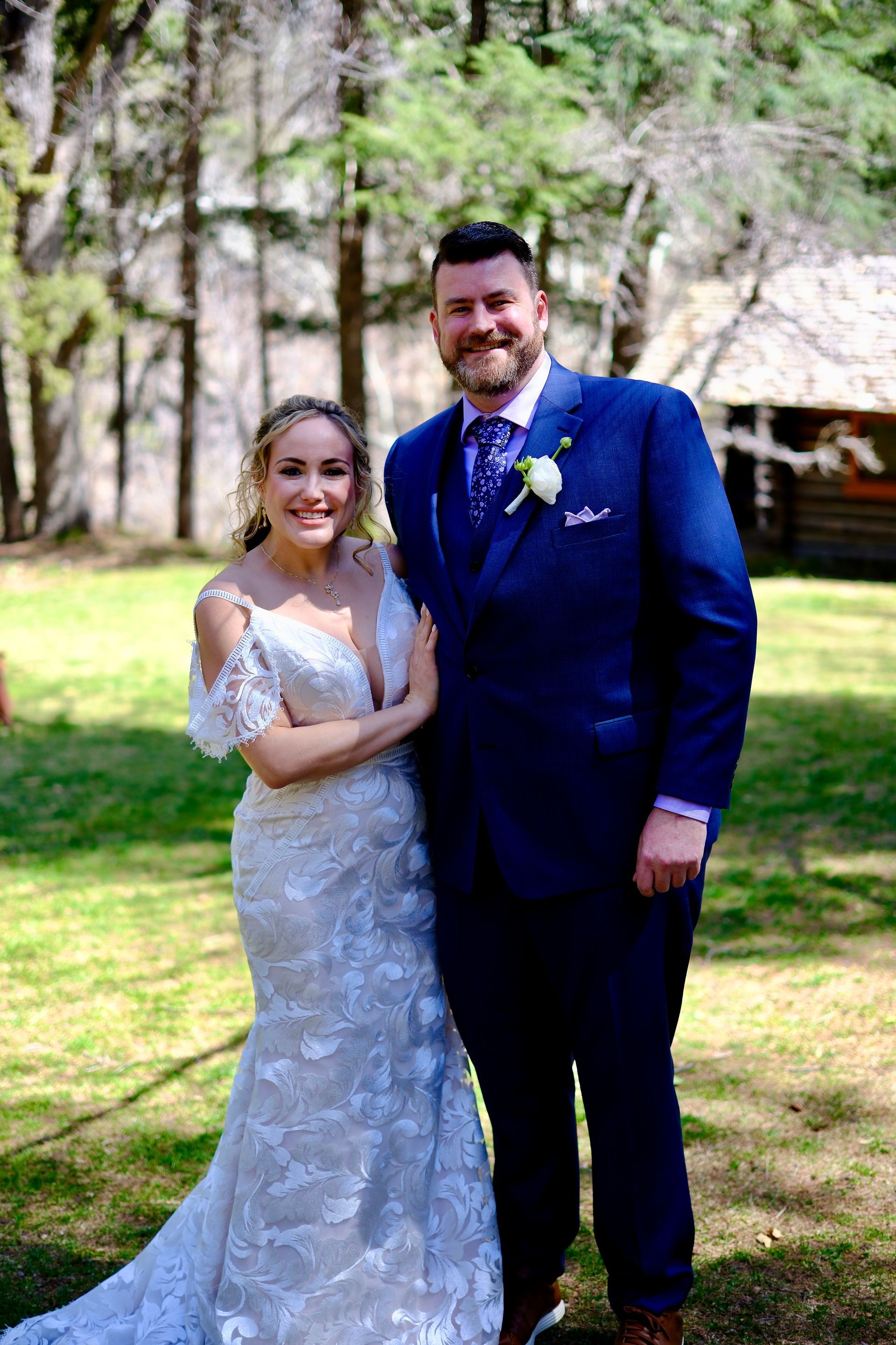 Wedding photo: smiling bride in white dress and groom in blue suit pose outdoors.