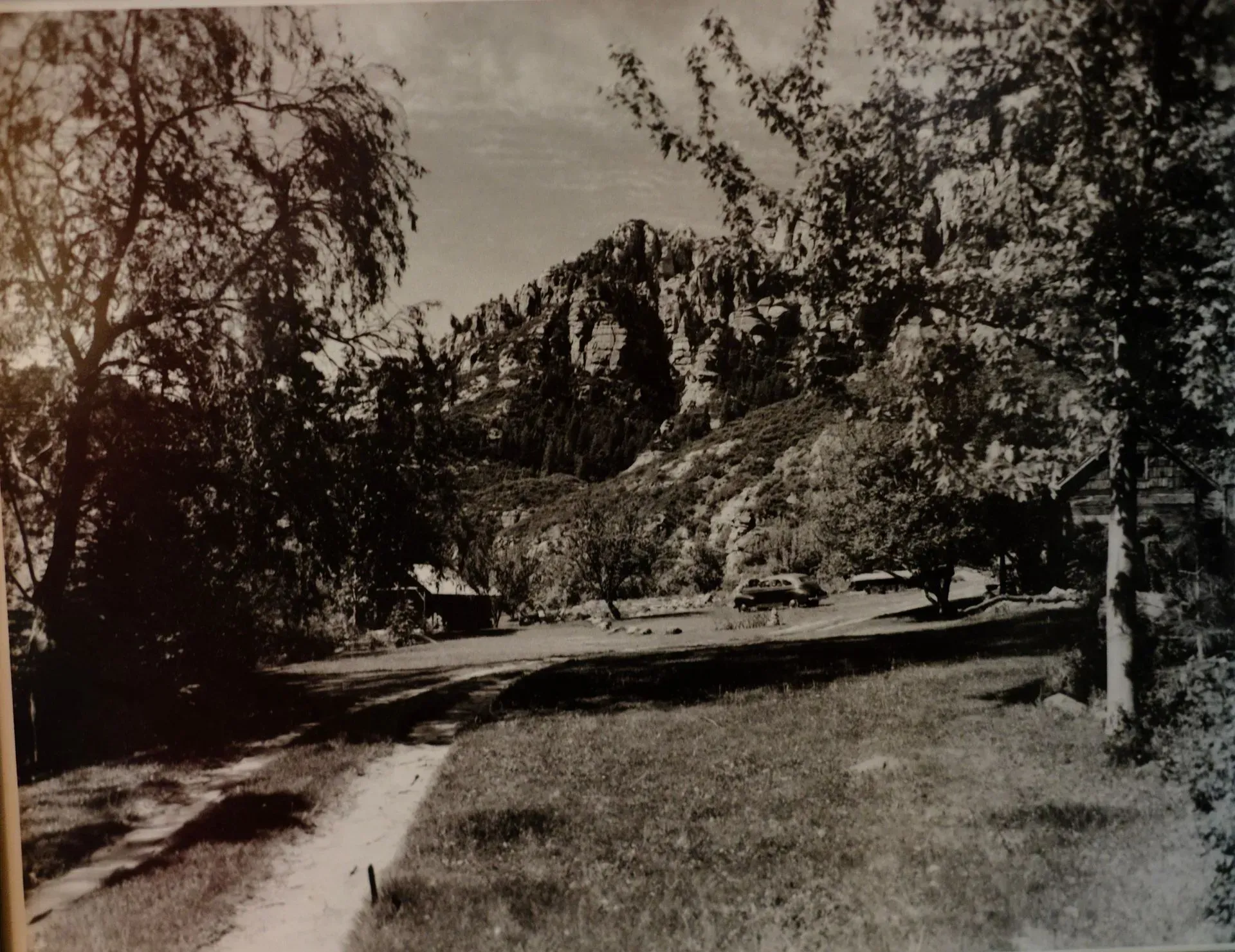 Black and white view of a mountain, trees, and cars parked in a grassy area with a dirt road.
