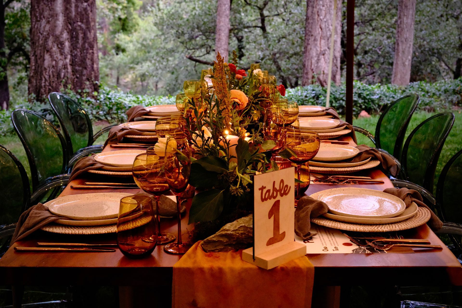 Formal dining table set in a forest with floral centerpiece, amber glasses, and green chairs. 
