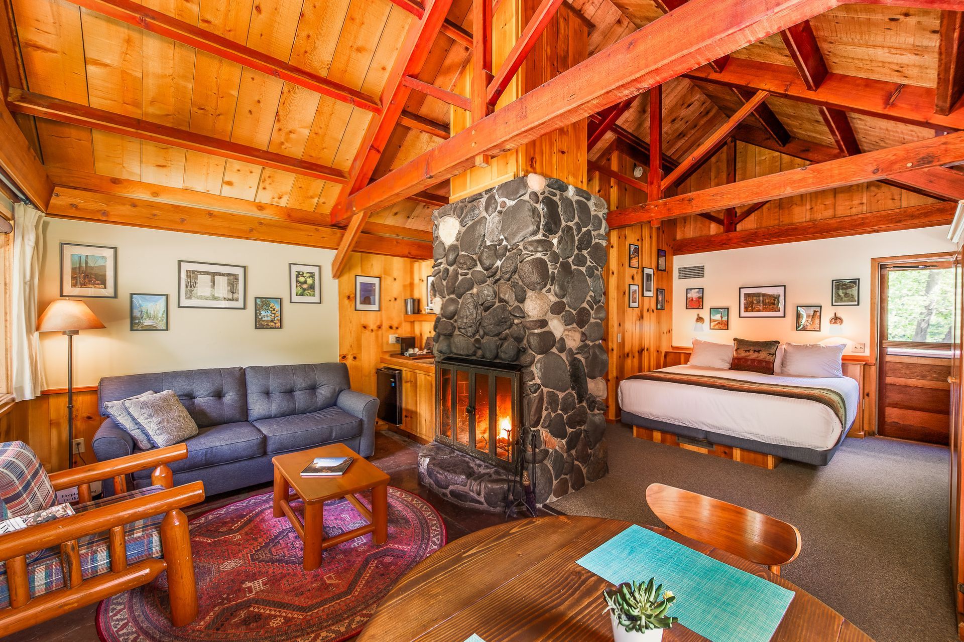 Cozy cabin interior with stone fireplace, wooden beams, a couch, bed, and dining table.