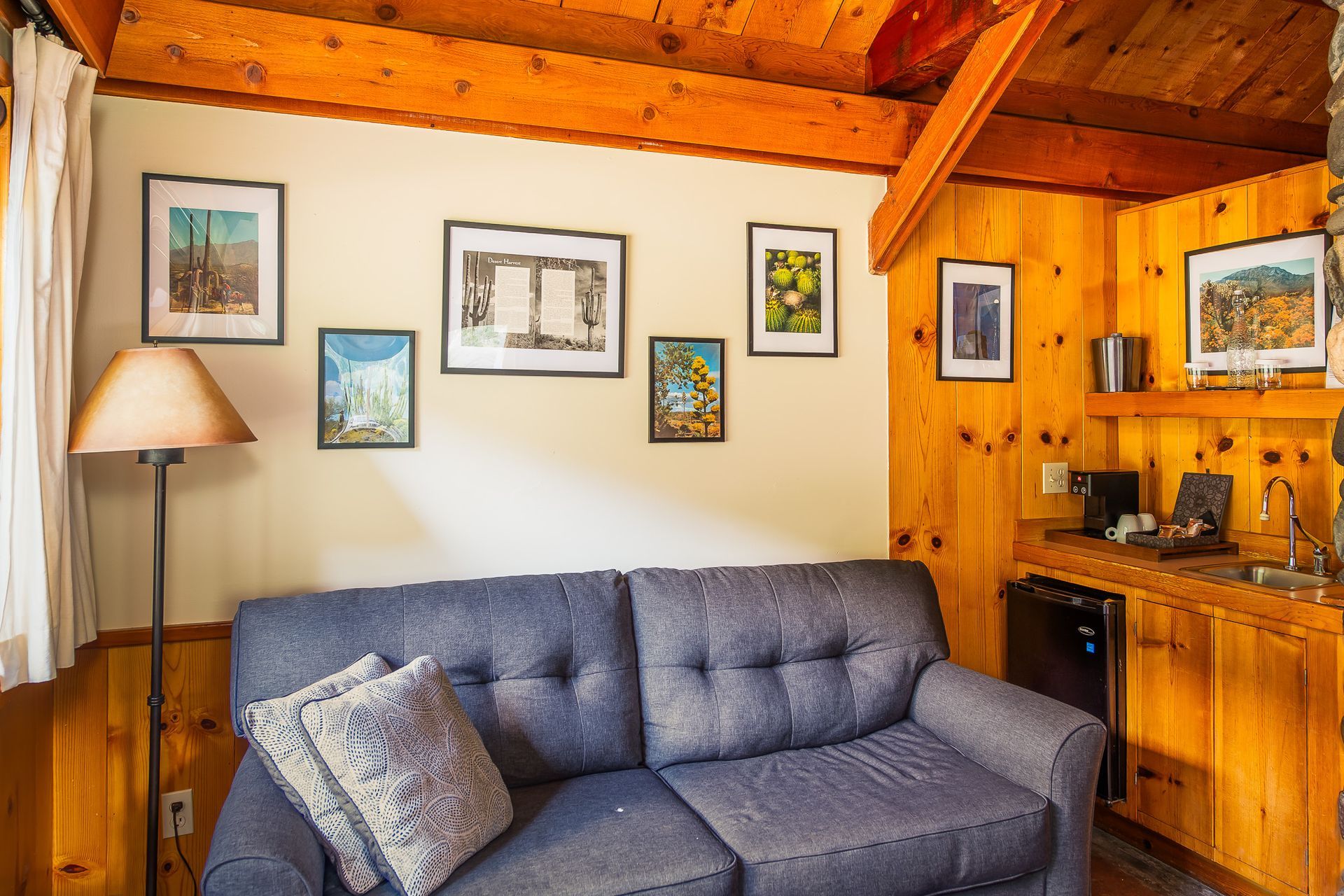 Cozy cabin interior with blue sofa, framed artwork, and wooden paneling. Includes a small kitchenette.