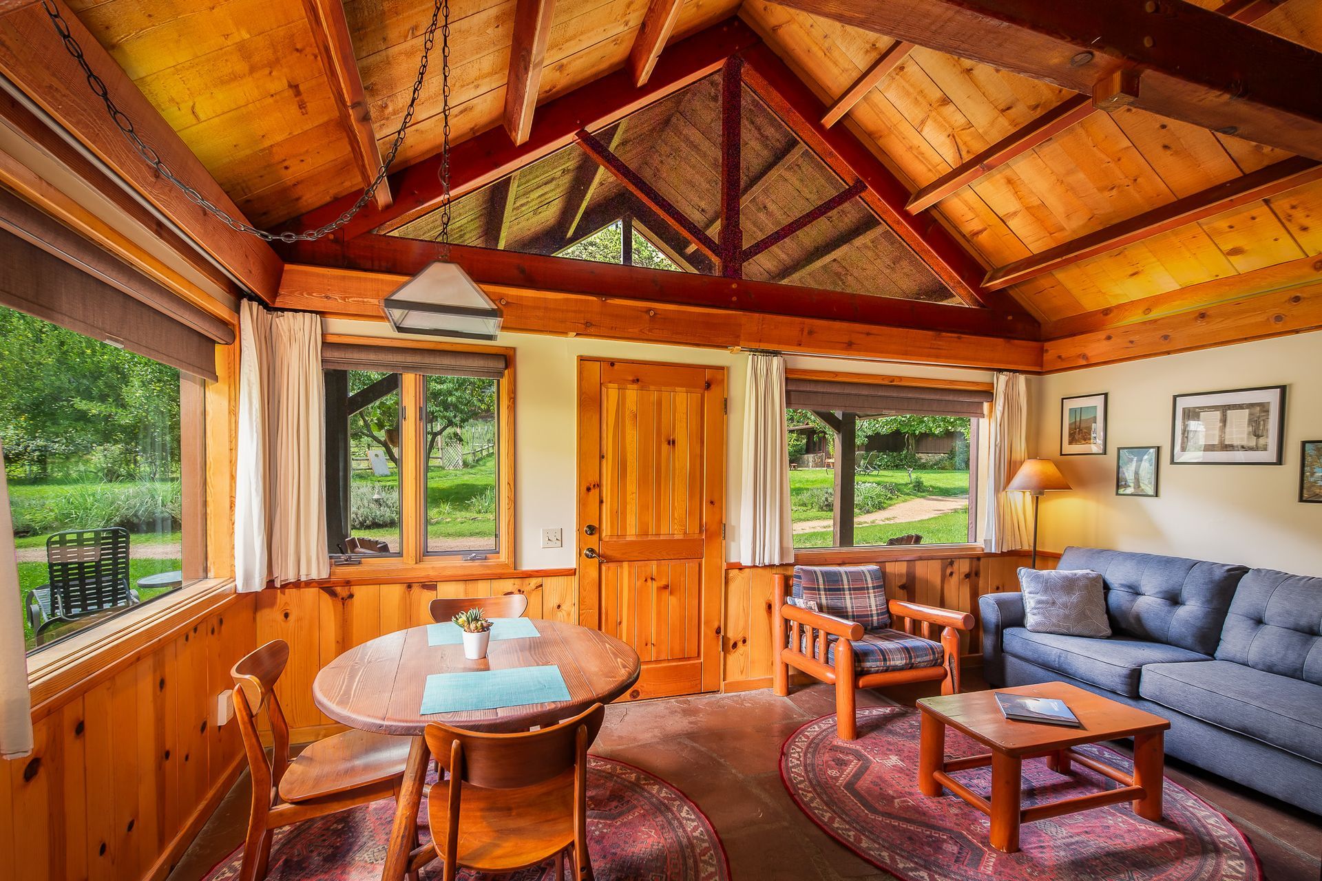 Cozy cabin interior with wooden walls, a dining table, sofa, and windows overlooking greenery.