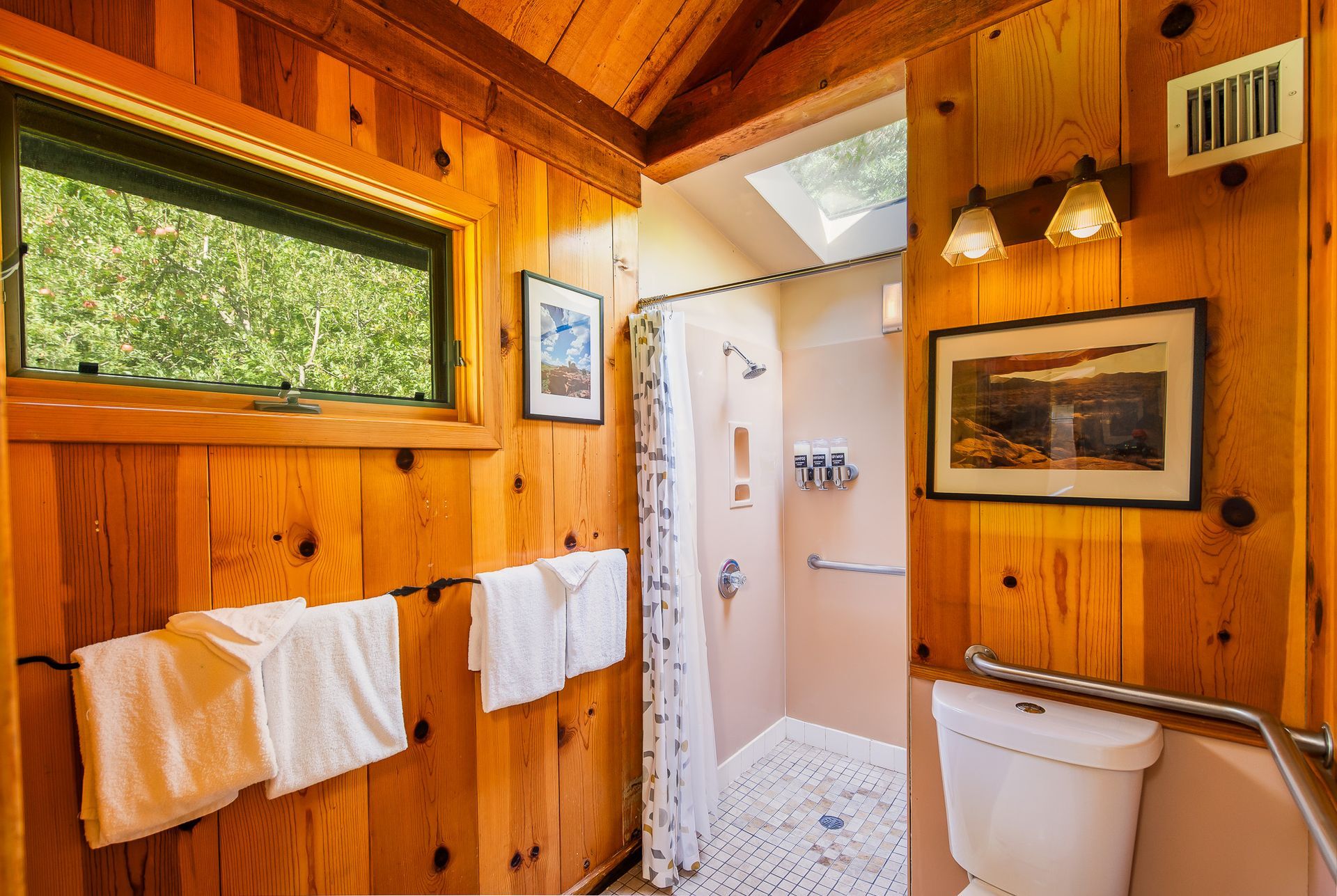 Rustic bathroom with wood paneling, shower, toilet, window with trees, and towels.