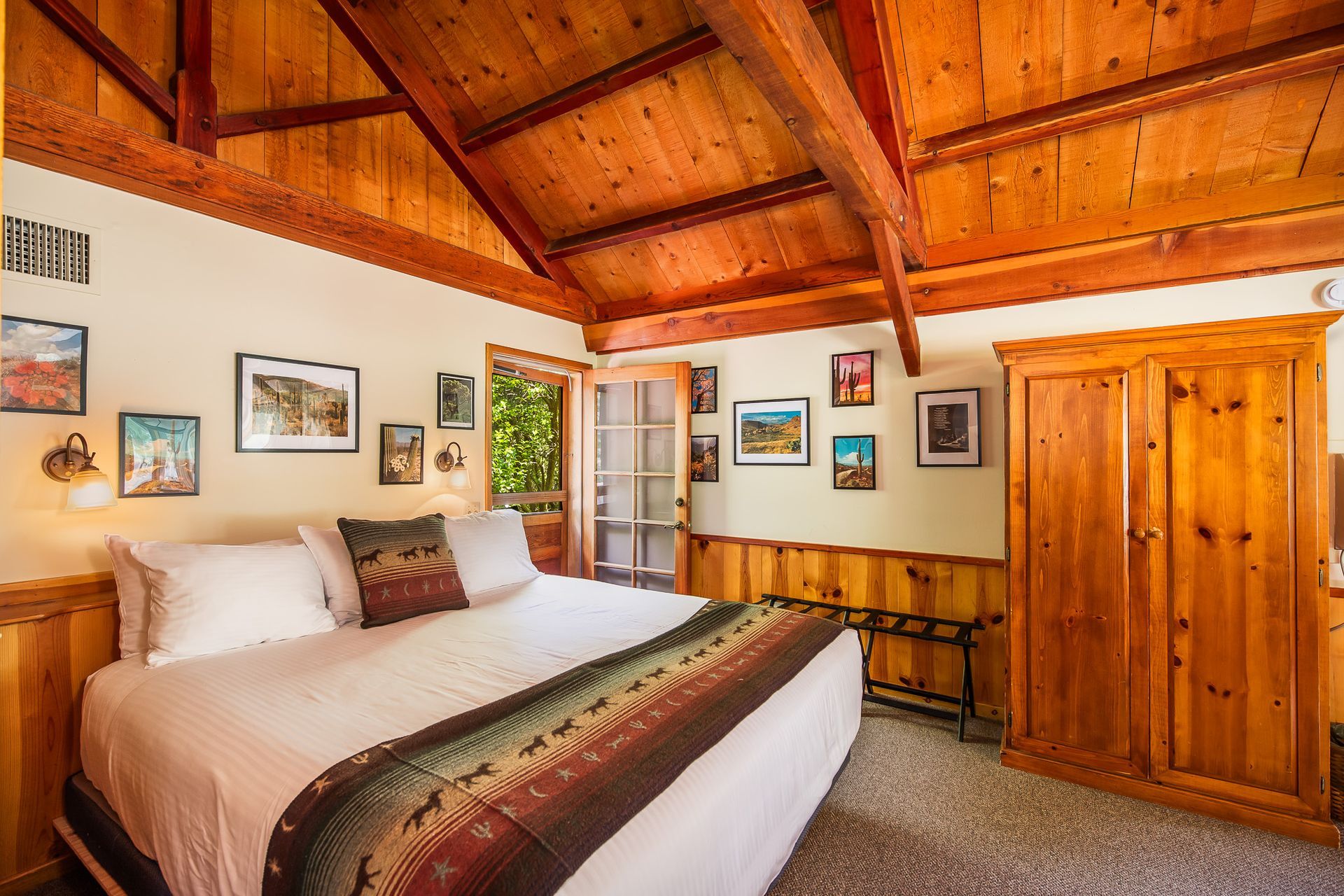Bedroom with wooden walls, ceiling, and wardrobe. Bed with colorful blanket, framed art on walls.