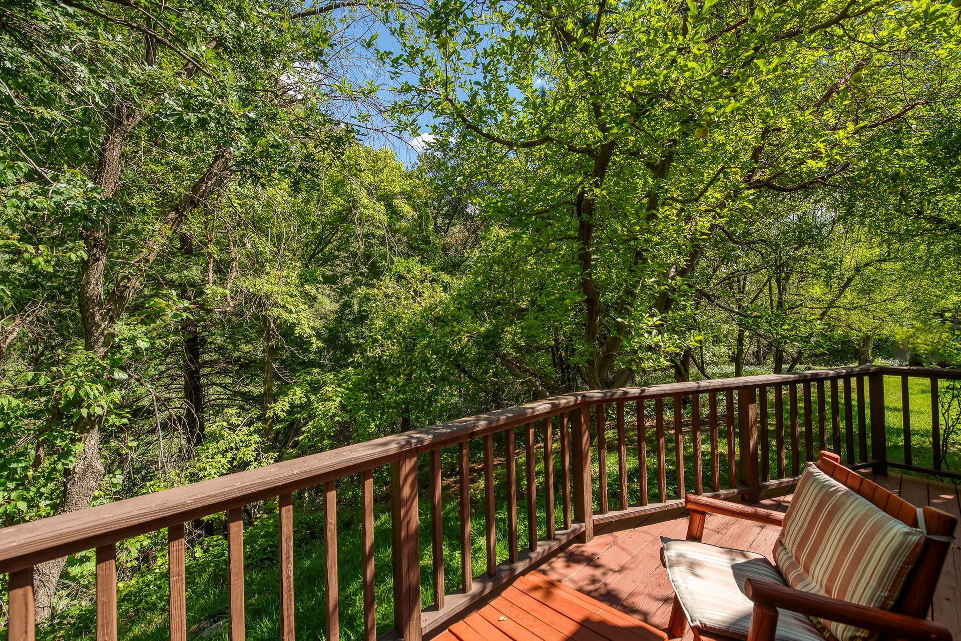 Wooden deck overlooking lush green trees. Brown railing, chair with patterned pillow.