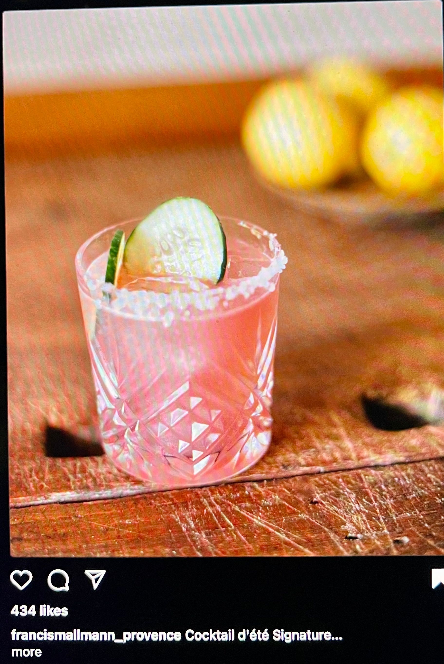 Pink cocktail in a patterned glass with a cucumber garnish, on a wooden surface, lemons in background.