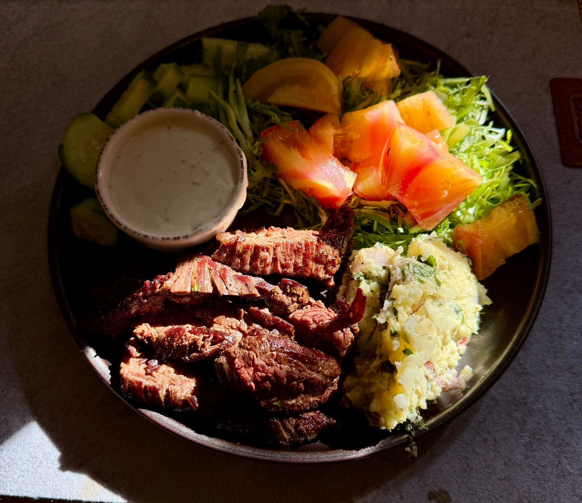 Grilled steak slices, salad with tomatoes and pineapple, potato salad, and creamy sauce on a plate.