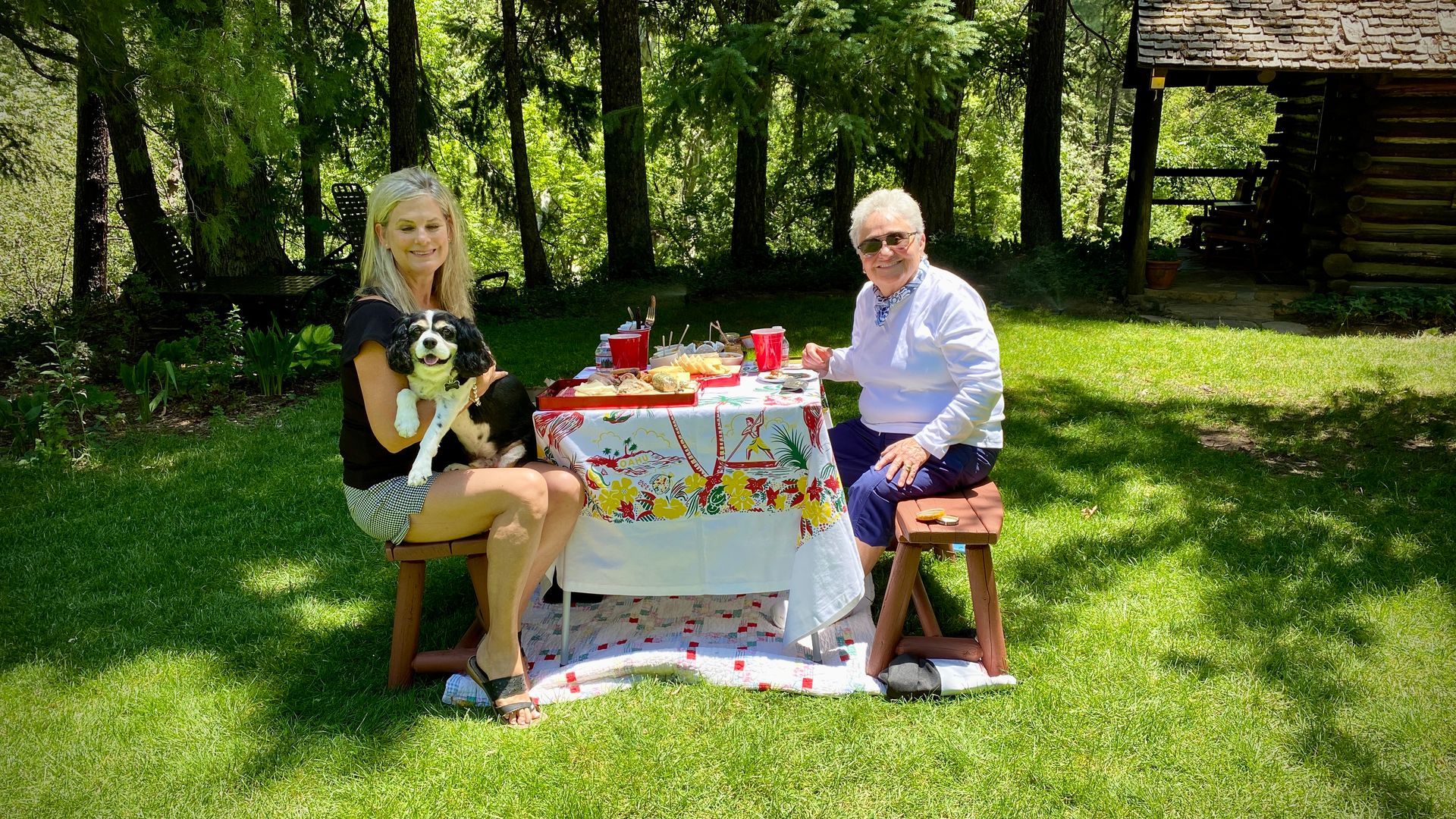 Woman holding dog, and older woman seated at picnic table in sunny garden.