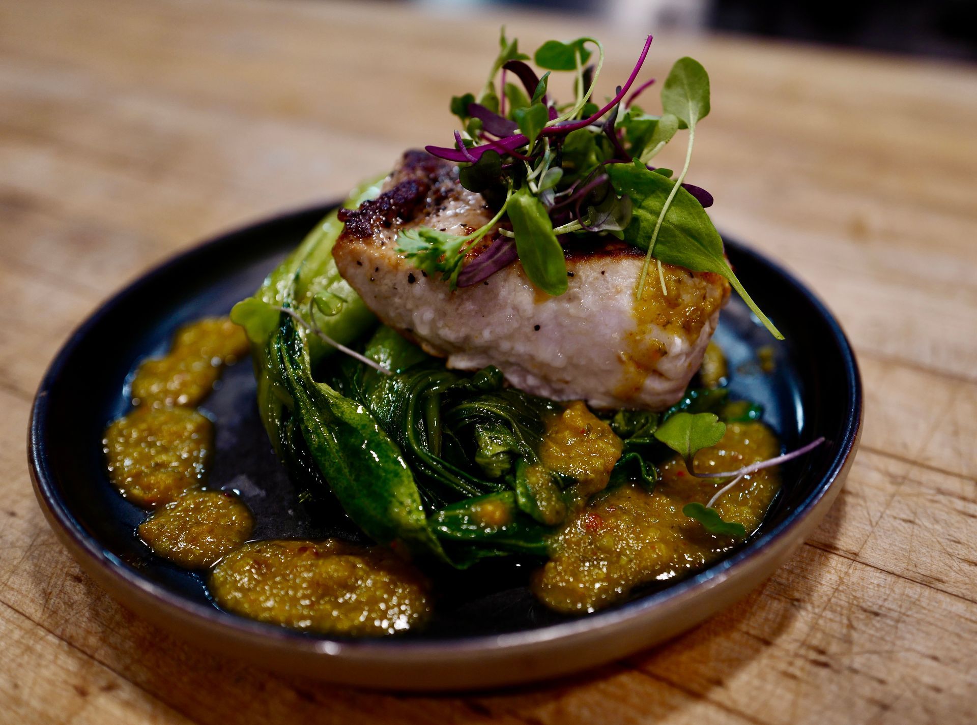 Seared fish atop sautéed greens, drizzled with sauce, garnished with microgreens on a dark plate, wooden surface.