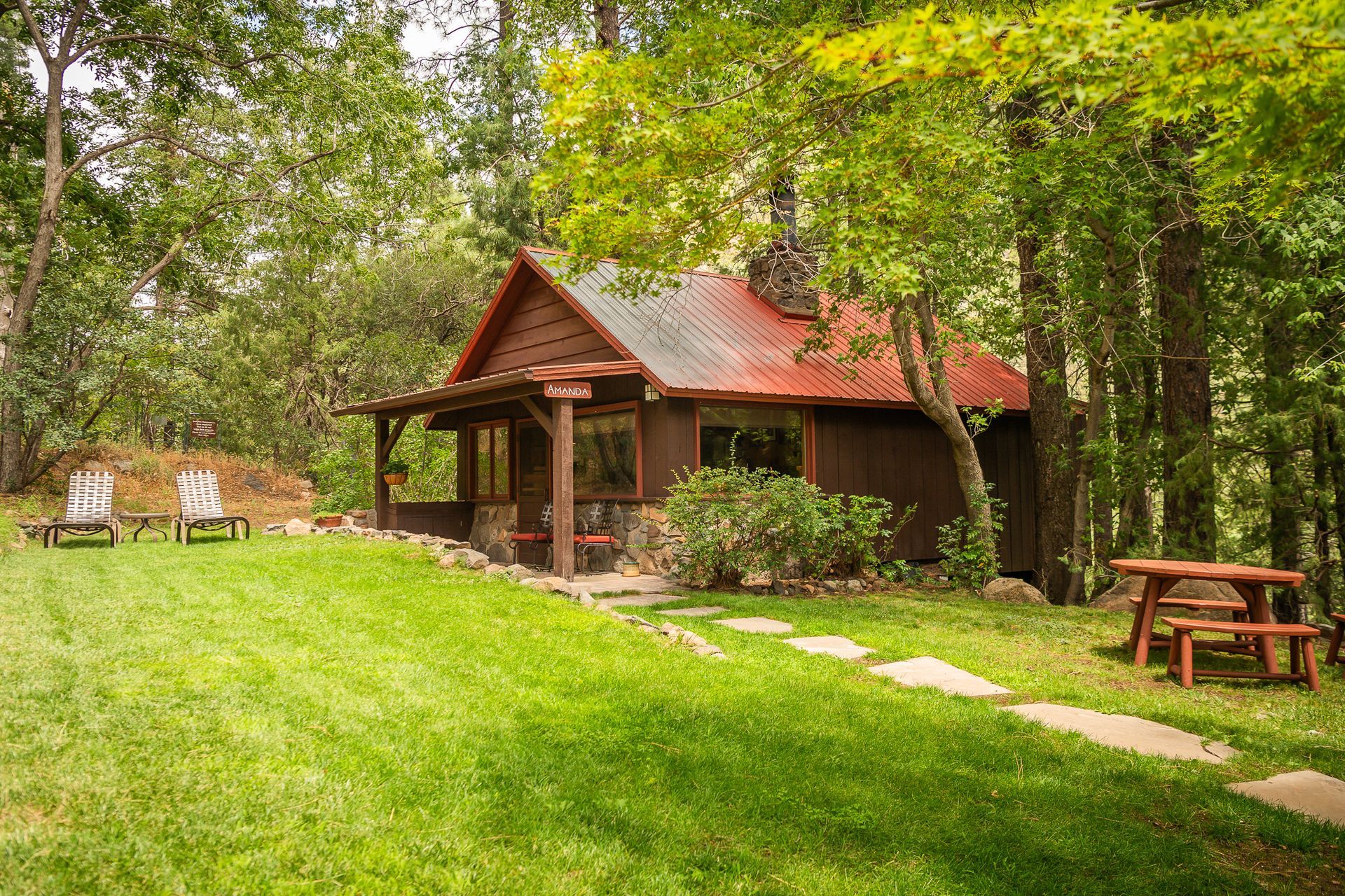 Cabin with red roof nestled in green forest, with lawn and picnic table.