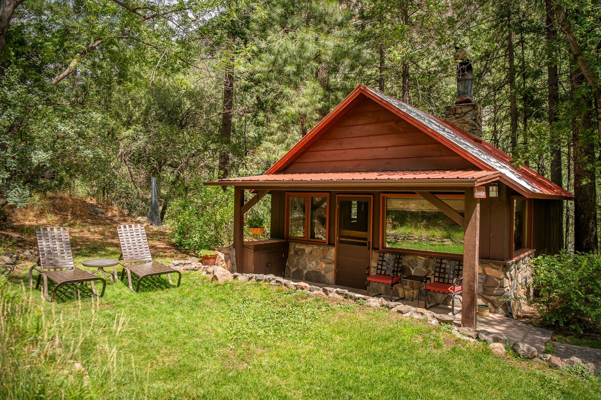 A rustic, brown cabin with a stone base, red roof, and chairs on a grassy lawn in a wooded area.