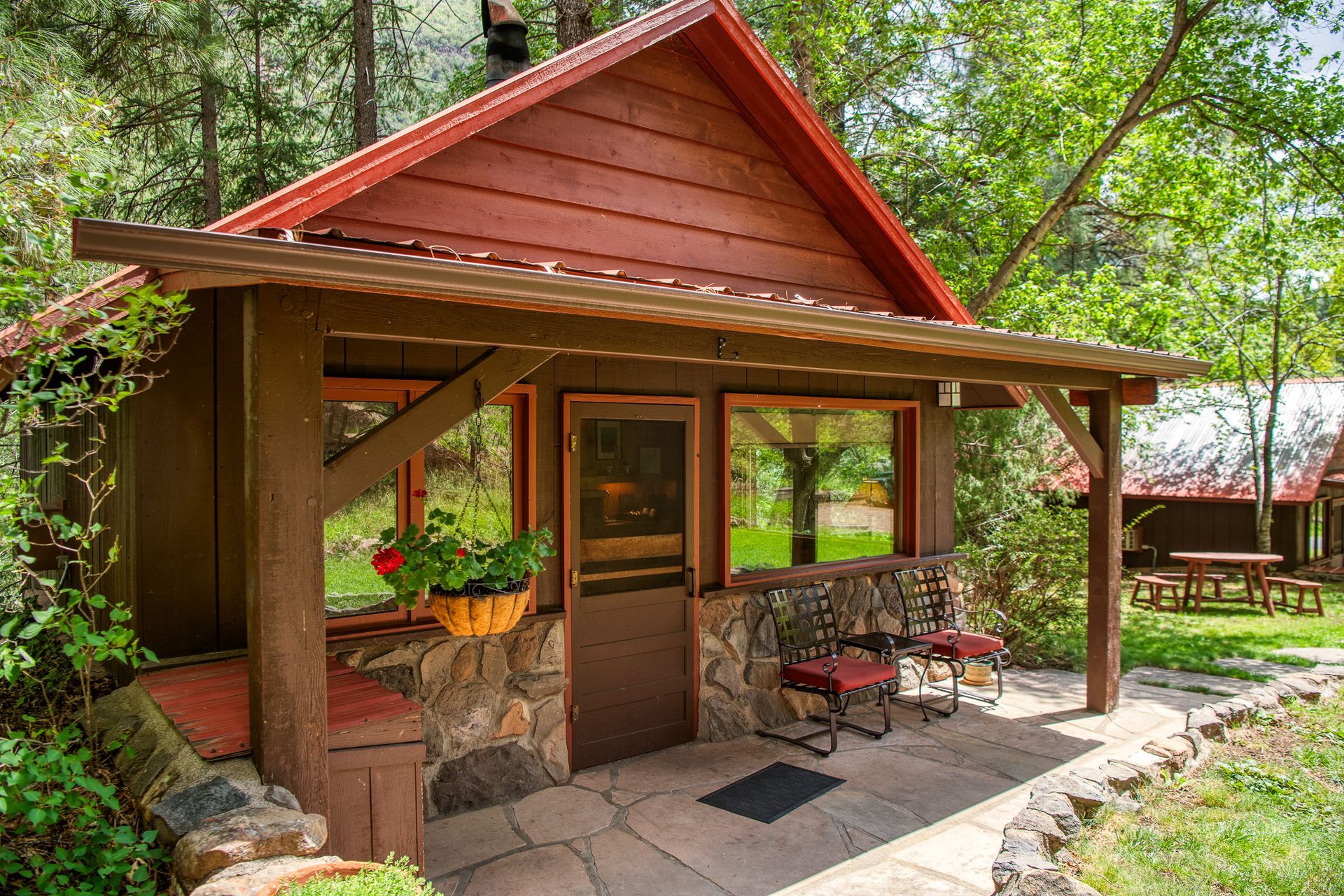Cabin with red roof, stone accents, and a covered porch with seating in a wooded setting.