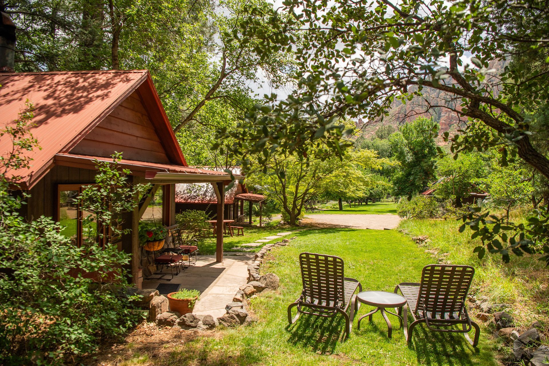 Wooden cabin with red roof nestled in lush green yard; two chairs and a table on the lawn.