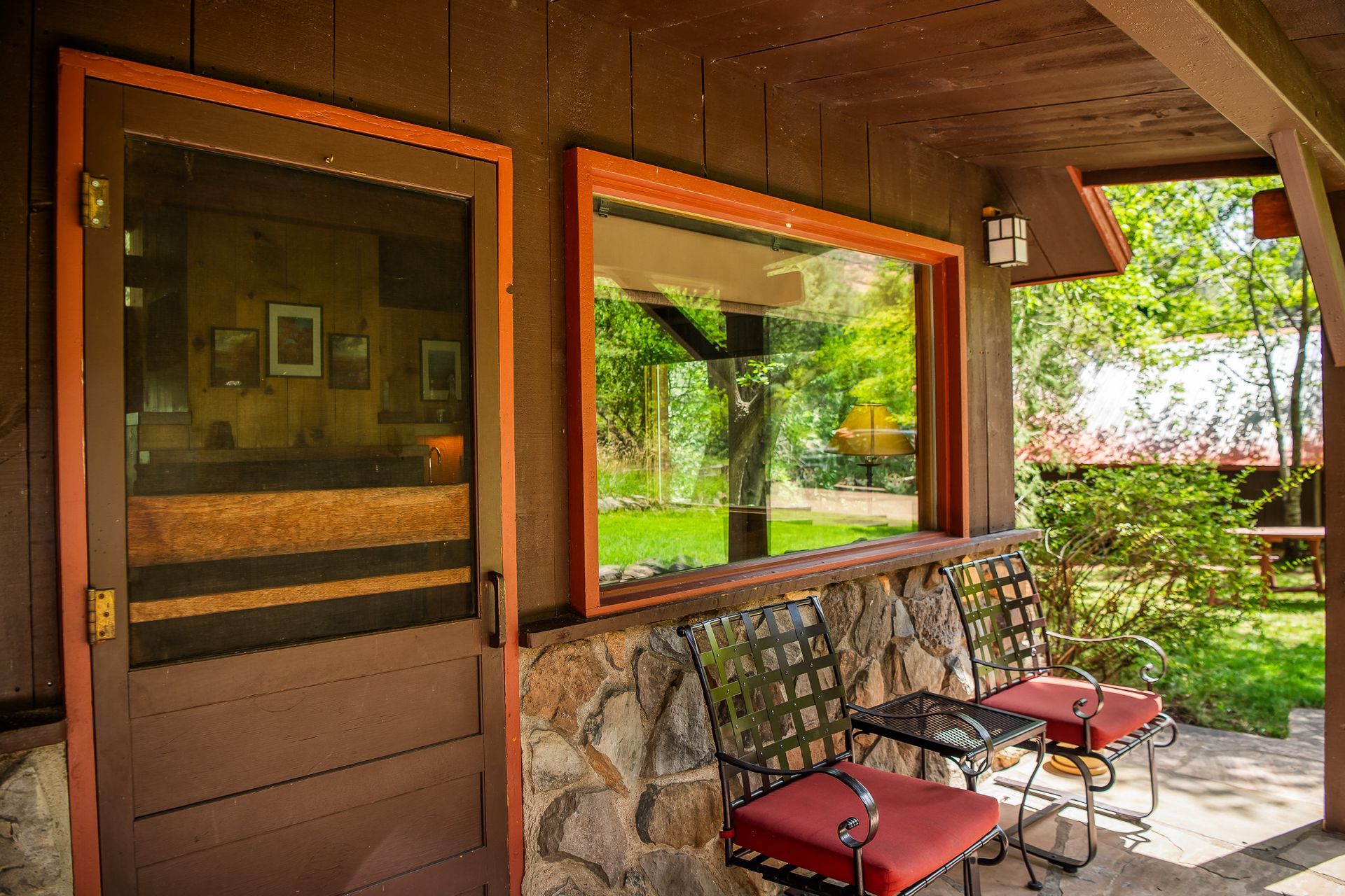 Brown cabin porch with screen door, window, and chairs with red cushions. Green trees and sunny outdoors.