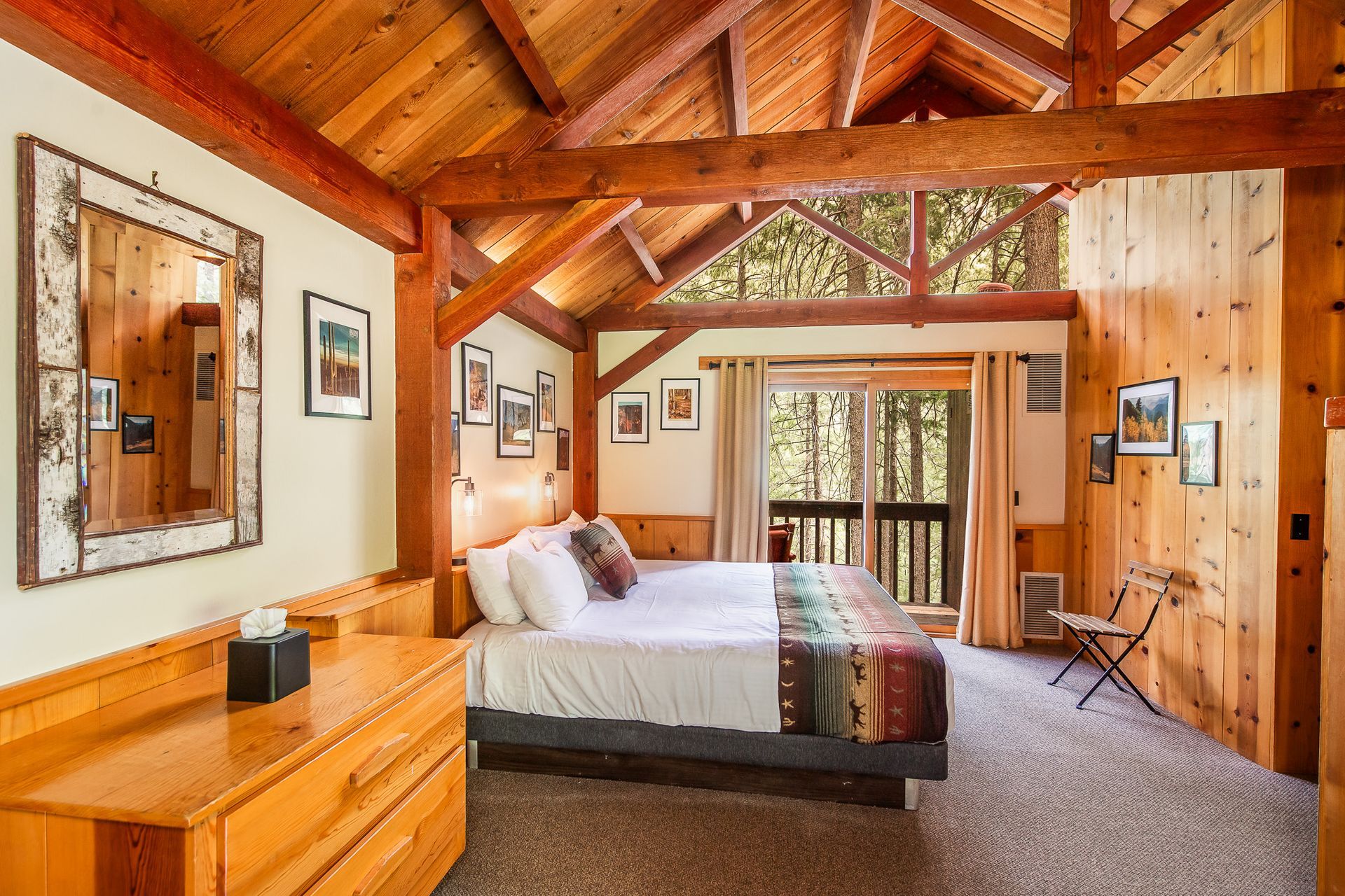Bedroom with wooden beams, a bed, dresser, and sliding door to a balcony overlooking trees.