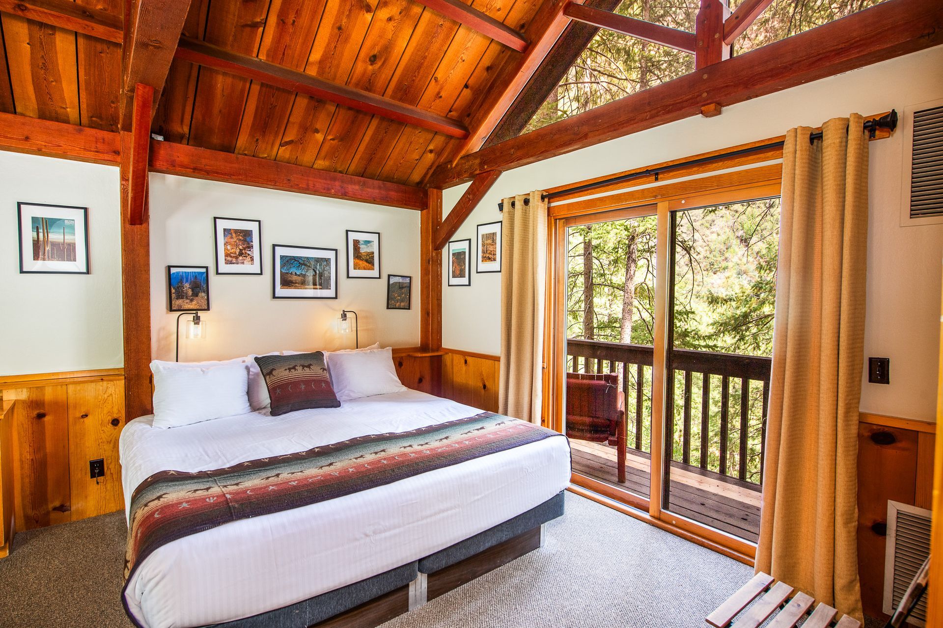 Bedroom with a king-sized bed, wooden beams, and balcony overlooking trees. Walls are light, and a rug covers the floor.