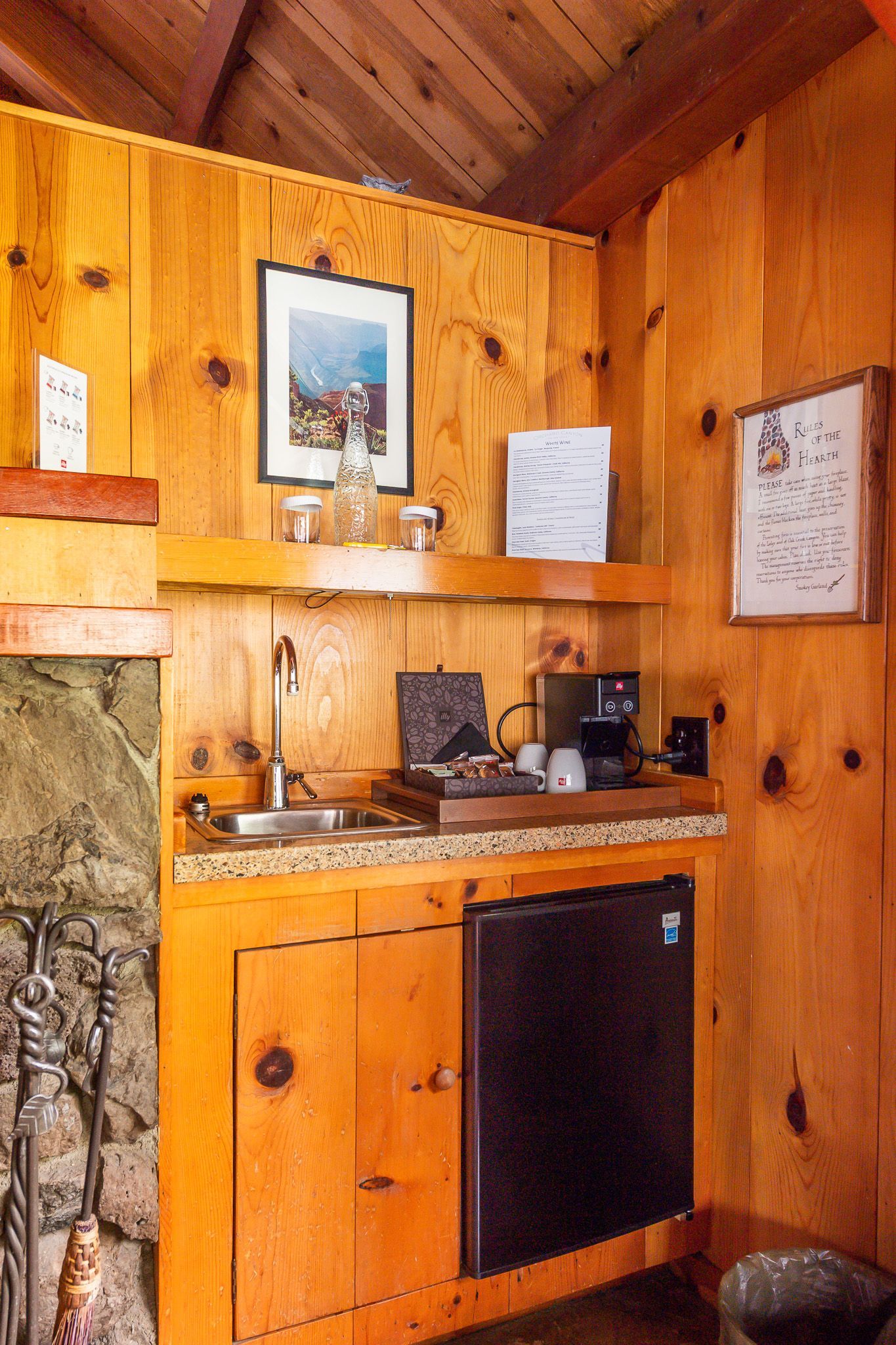 Small kitchenette with wood paneling. Includes a sink, mini-fridge, coffee maker, and shelves.