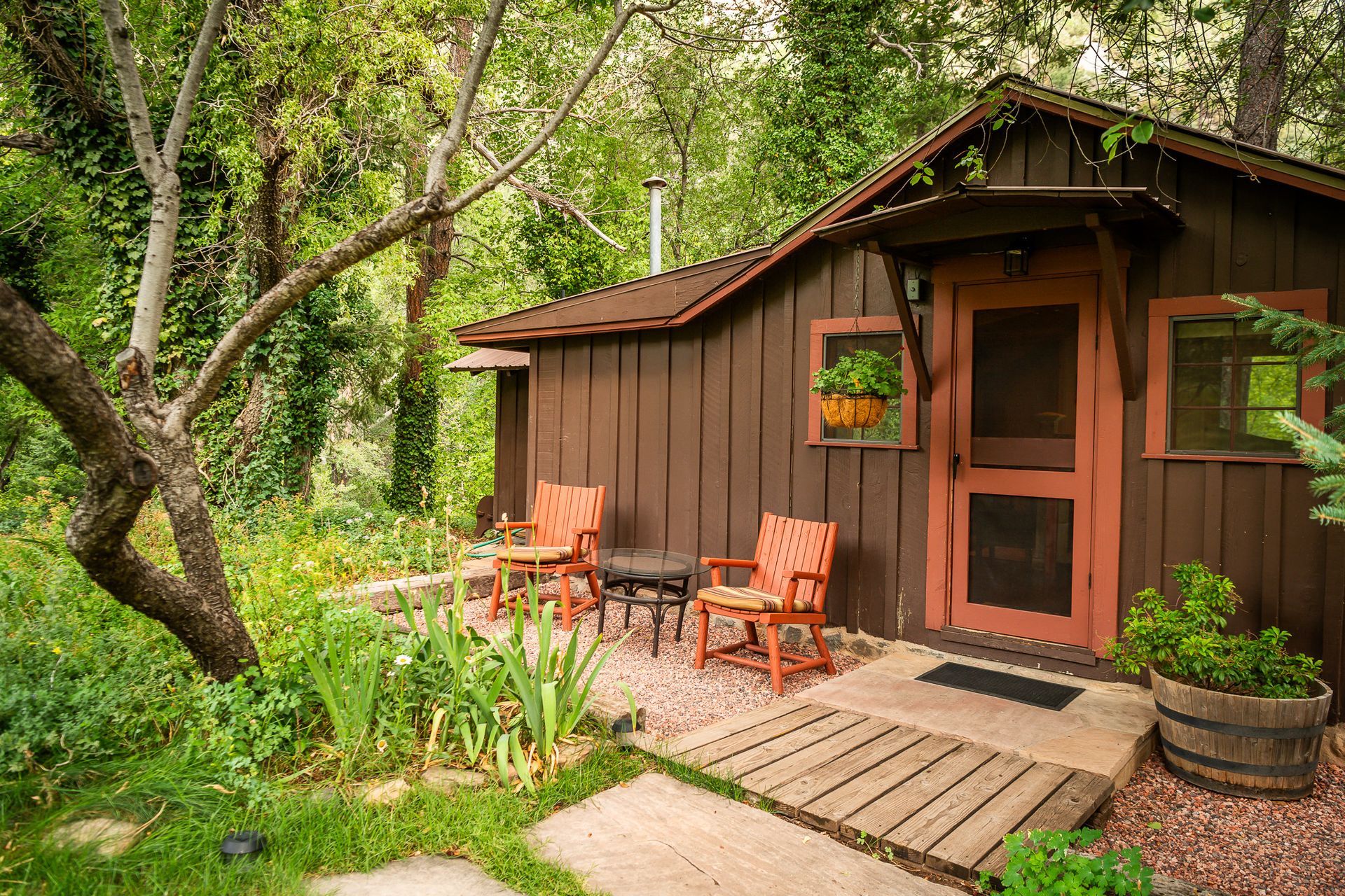 Cozy brown cabin with seating area on a stone patio surrounded by lush greenery.