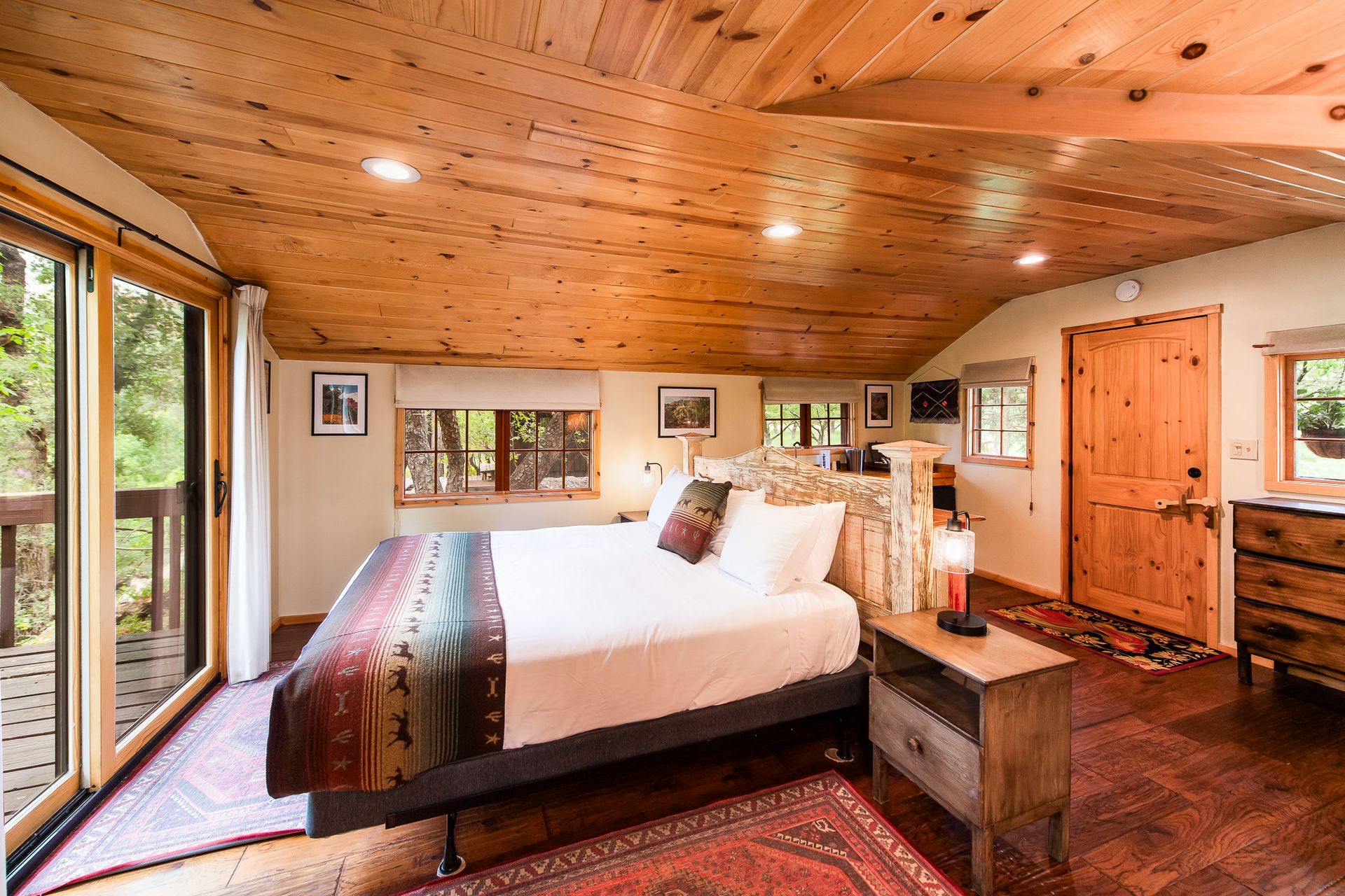 Bedroom with wood ceiling and walls, bed with colorful blanket, balcony access.