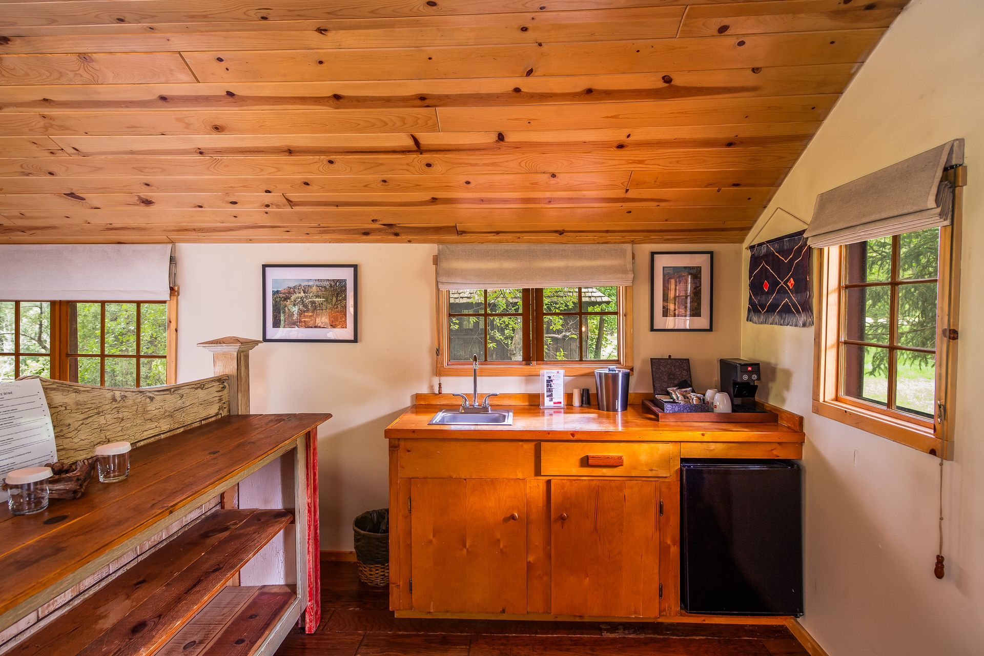 A rustic kitchenette in a room with a wooden ceiling and small windows. Features a mini fridge, sink, and coffee maker.