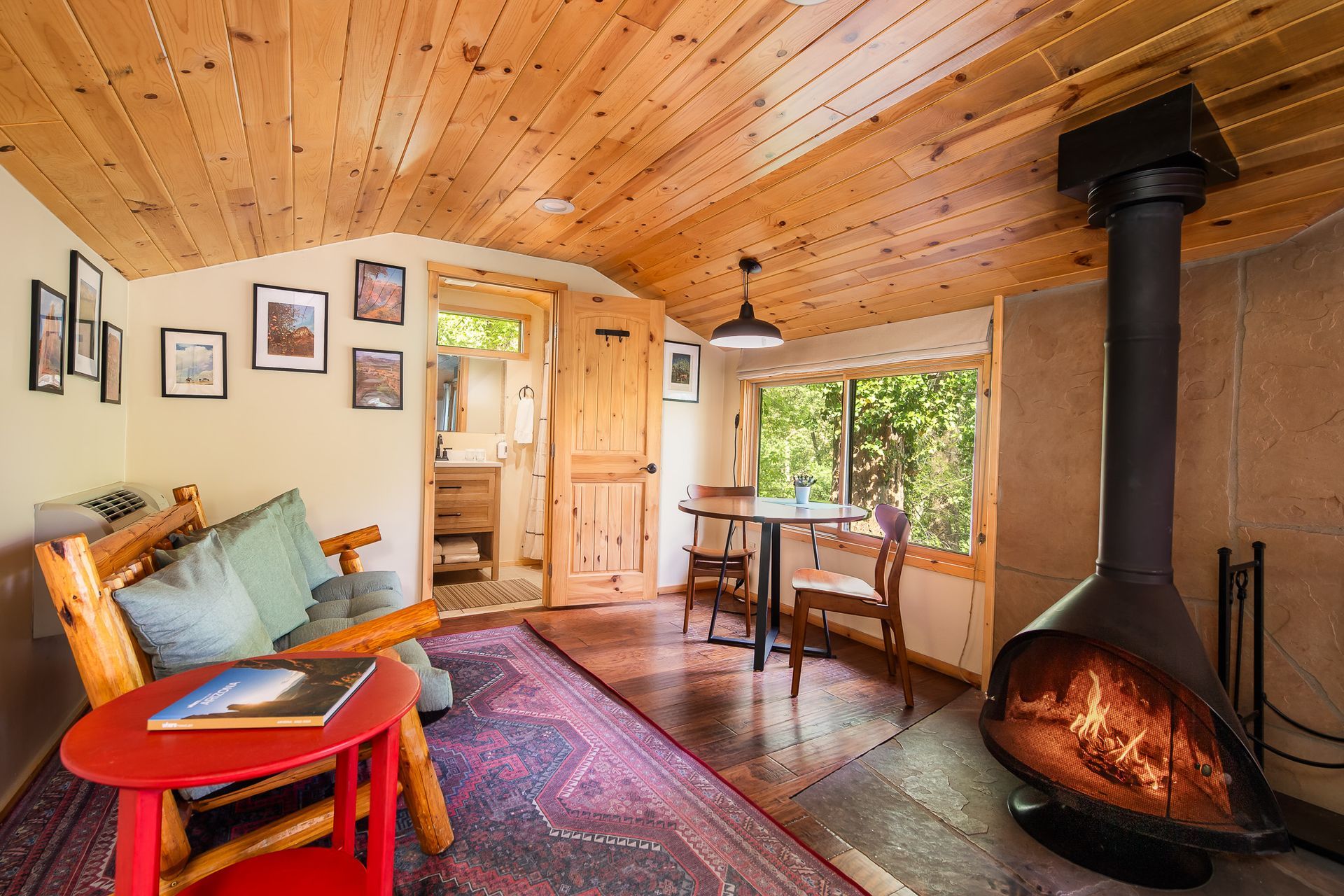 Cozy cabin interior: fireplace, wood ceiling, small dining table, sofa, artwork, doorway to bathroom.