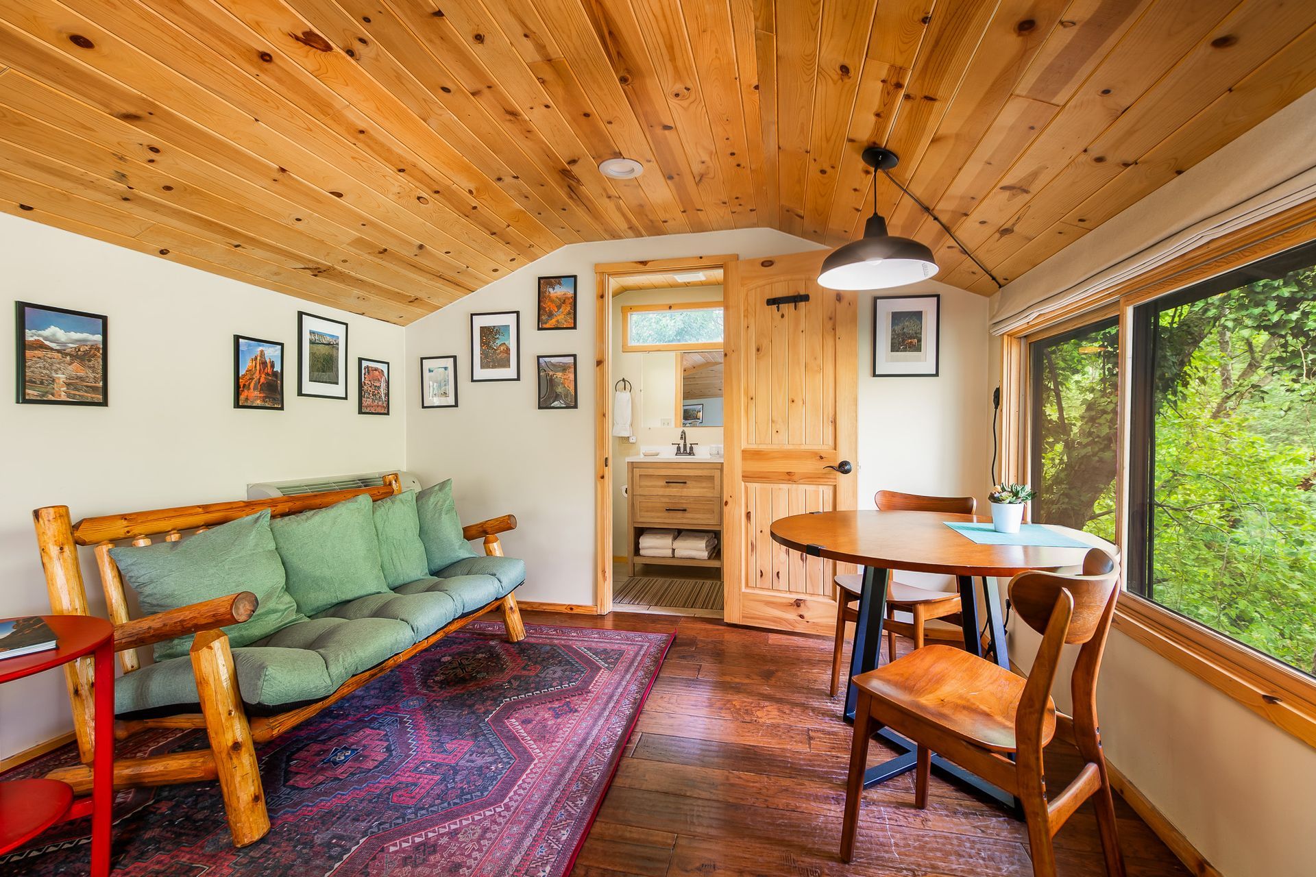 Cozy cabin interior with wood ceiling, sofa, round table, framed art, and a view of greenery.