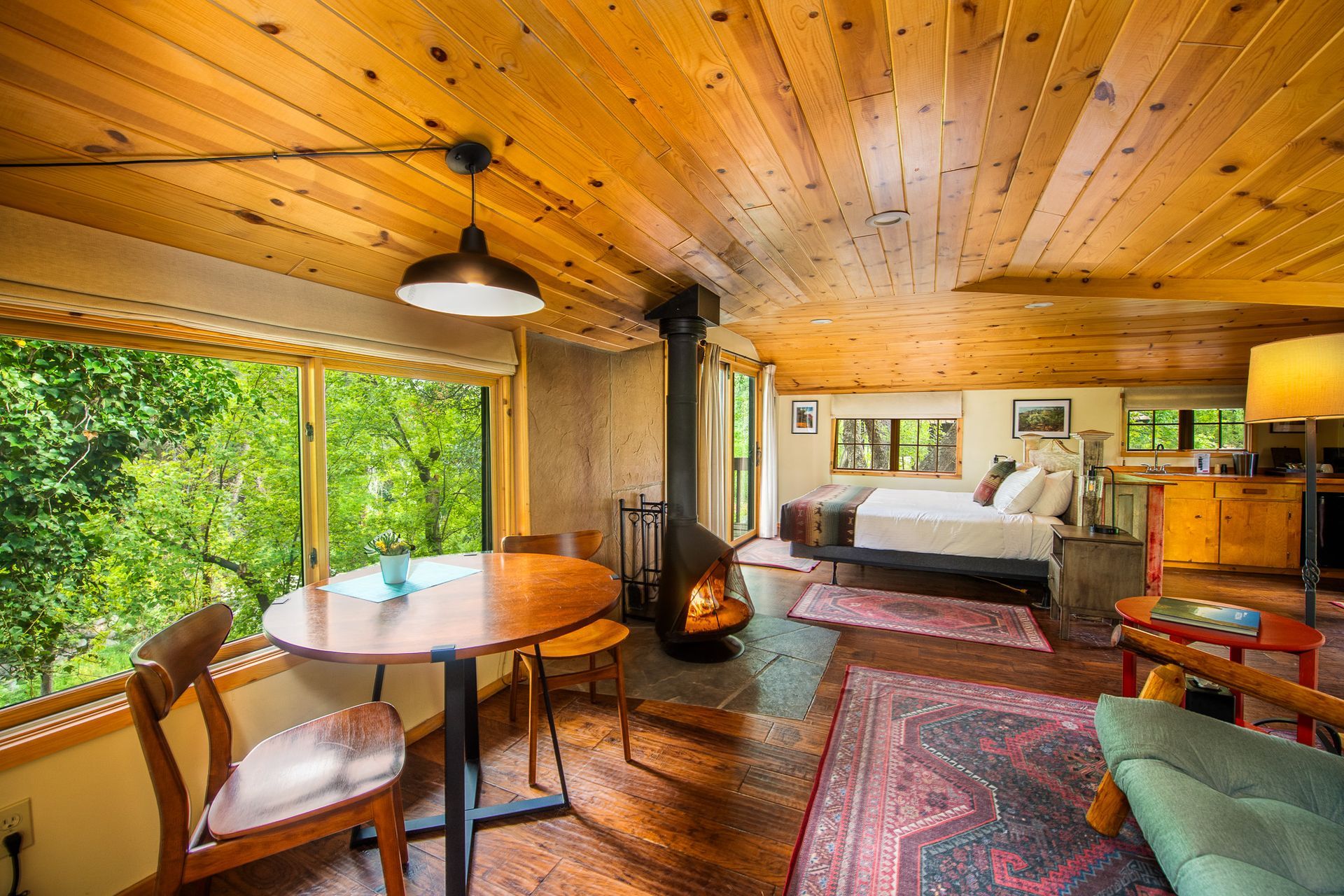 Cozy cabin interior with wood ceiling, large windows, fireplace, dining area, and bed.