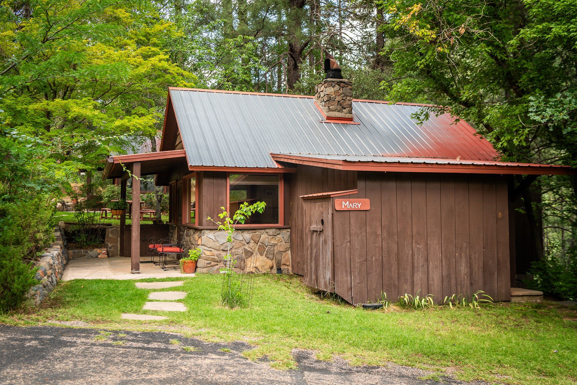 Cabin with brown siding and a stone chimney nestled in a wooded area.