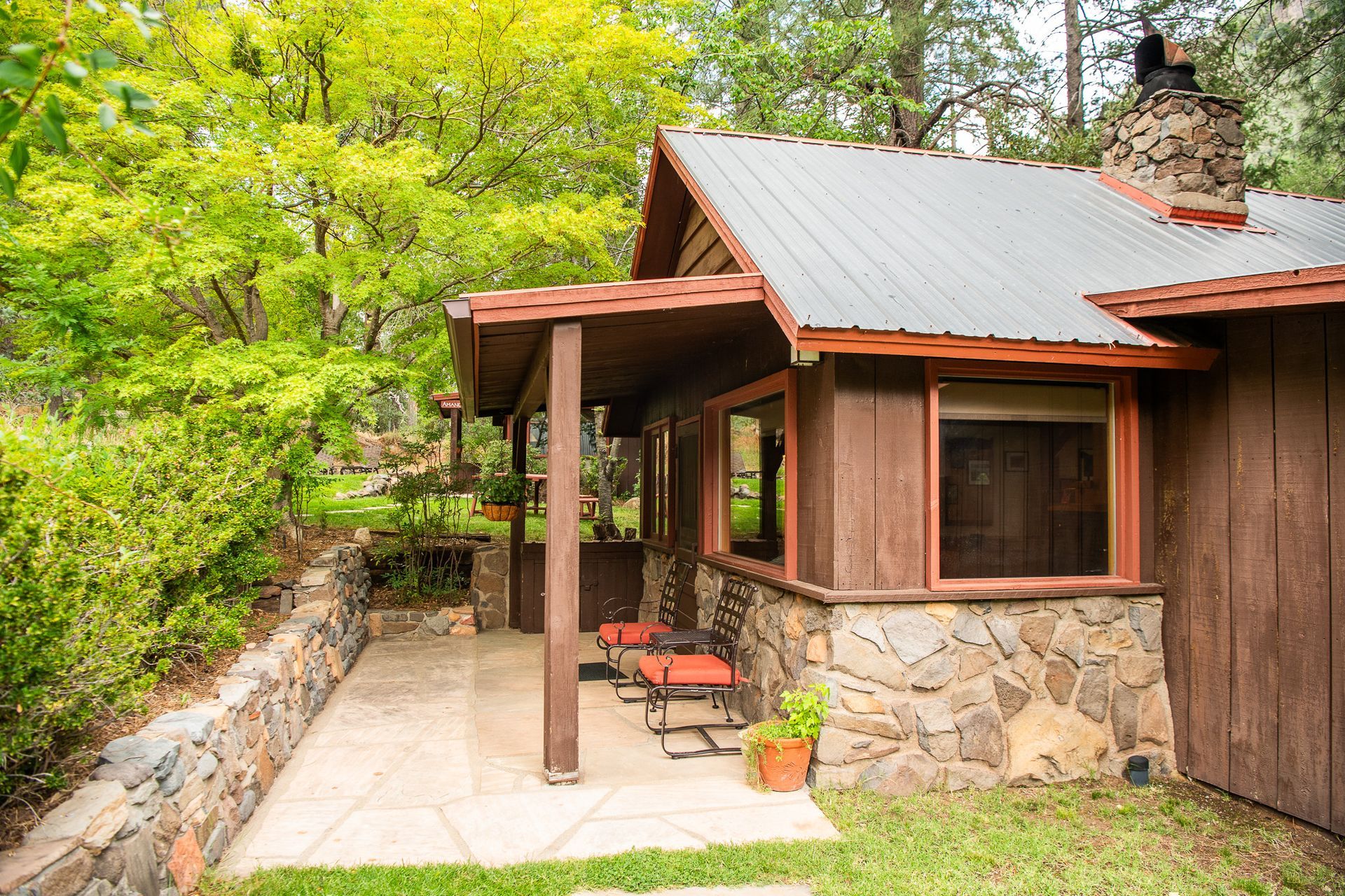 Rustic brown cabin with stone accents, porch, and metal roof surrounded by lush greenery.
