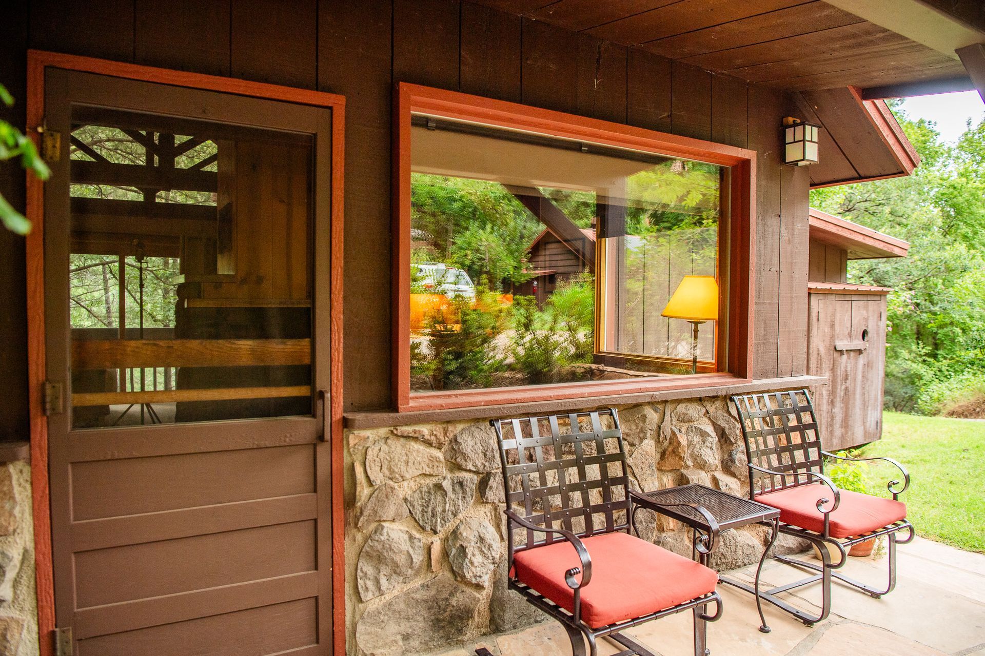 Porch with a stone base, brown siding, red-cushioned chairs, and a view of trees through a large window.