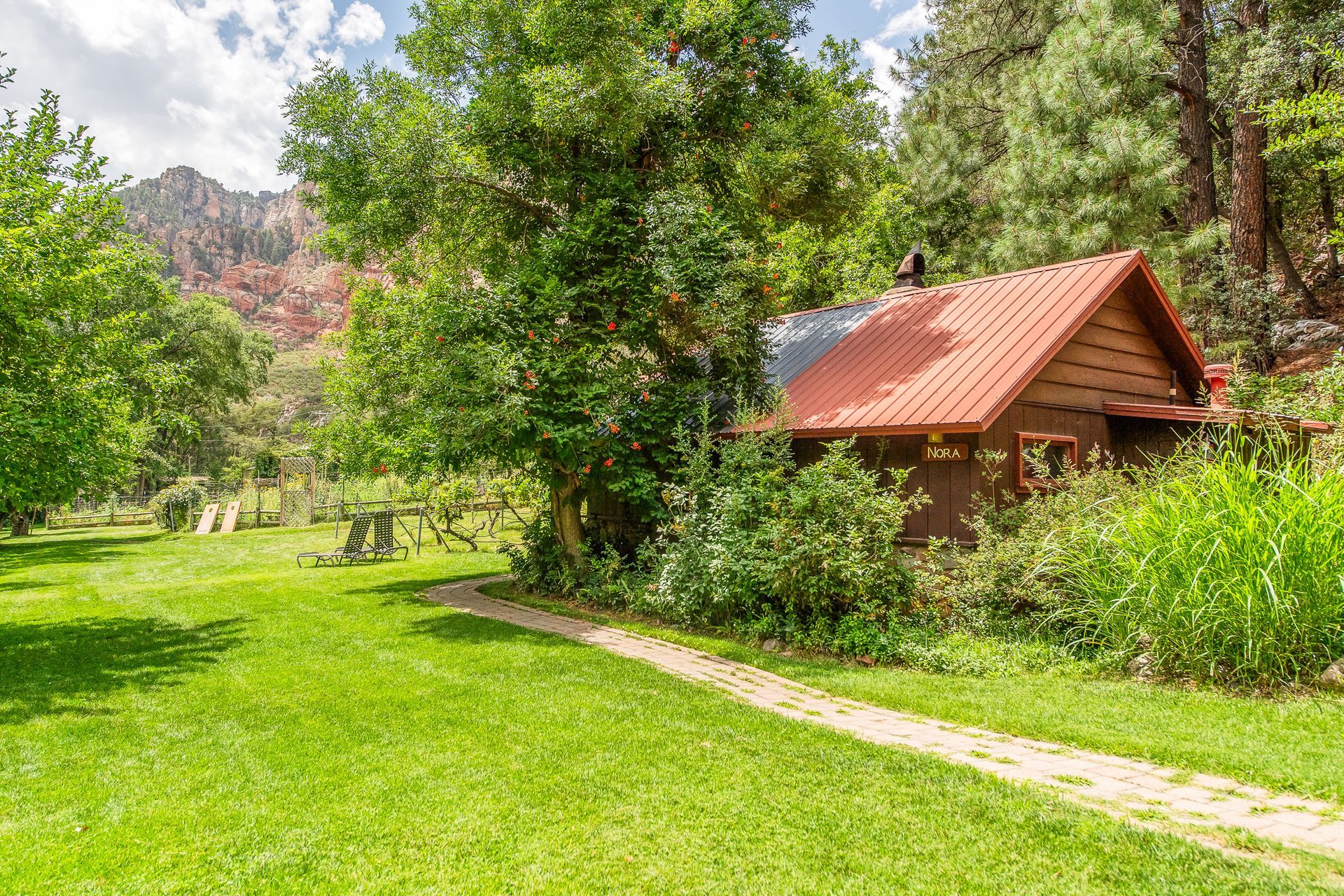 Cabin with red roof nestled in green grass, trees, and path leading to it.