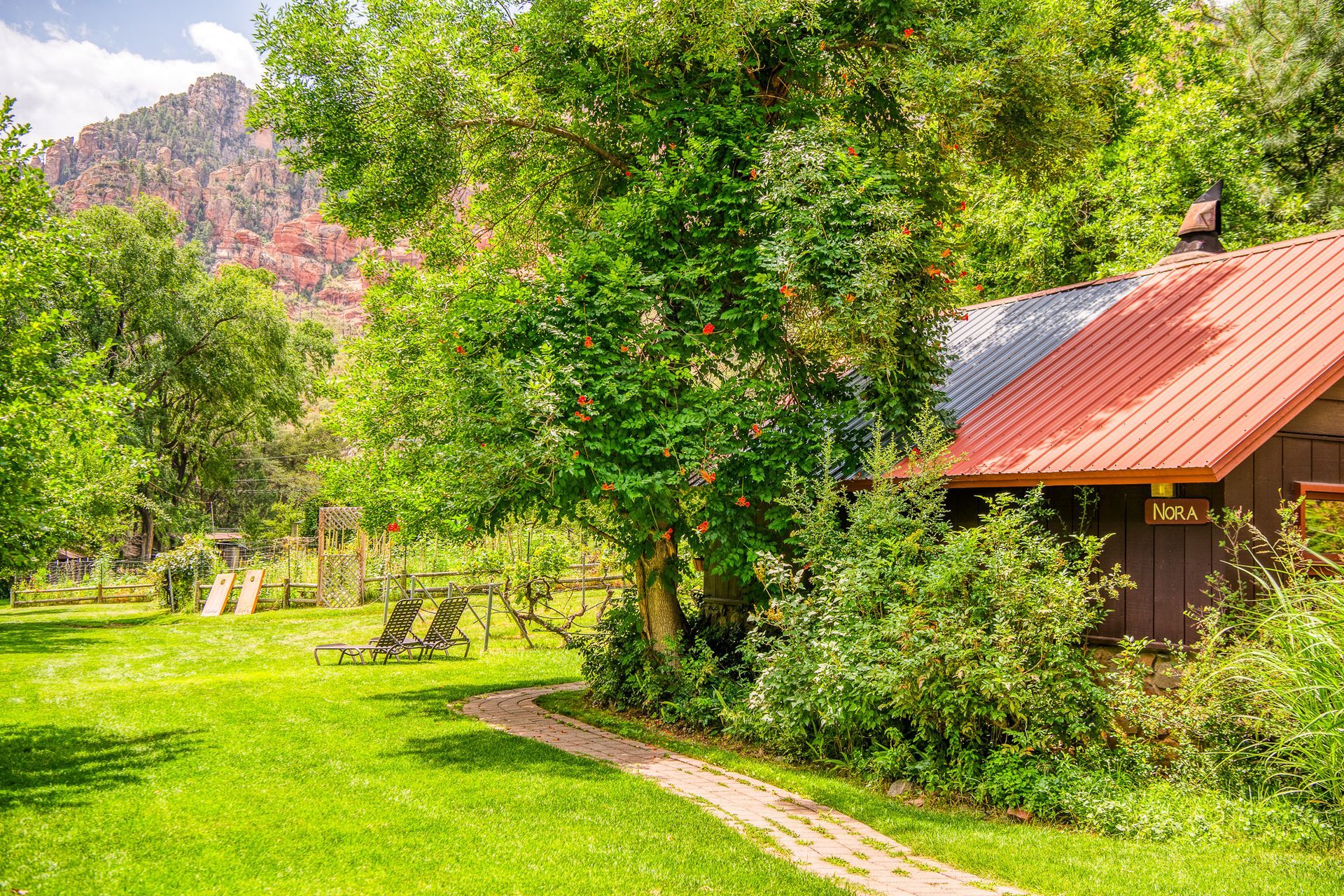 Lush green yard with a stone path leading to a cabin under a tree, mountains in the background.