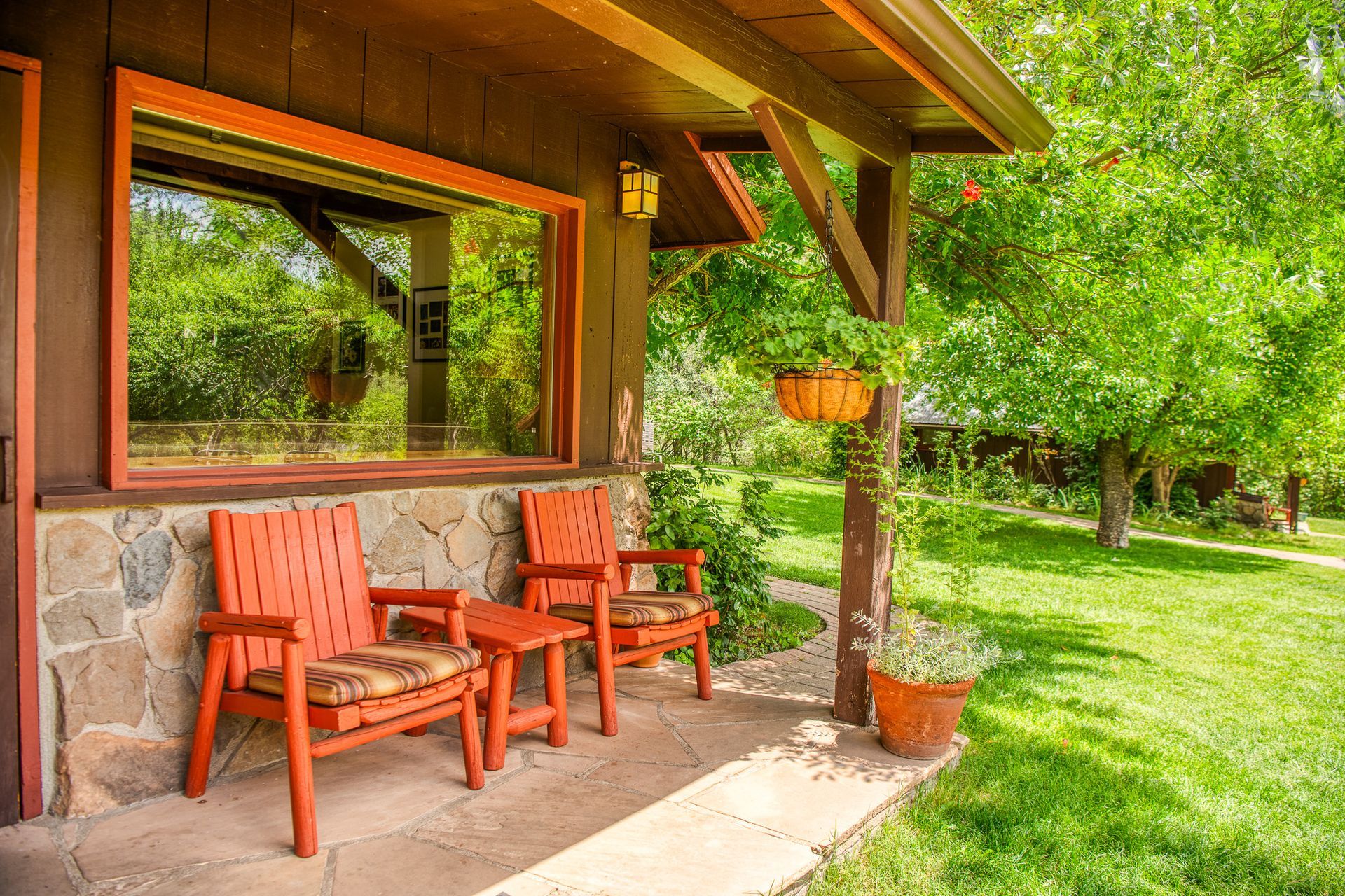 Two orange chairs and a small table on a porch, set against a stone wall and a lush green yard.