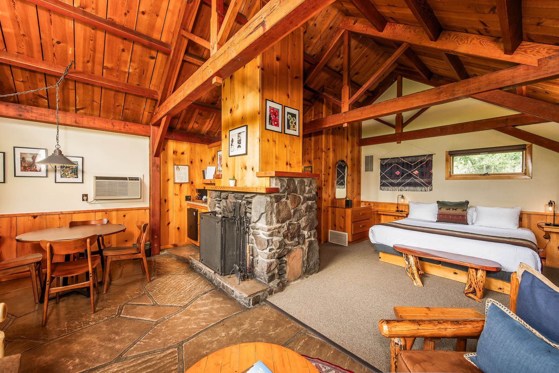 Cozy cabin interior with wood paneling, stone fireplace, and a bed. Furniture includes a table, chairs, and a love seat.