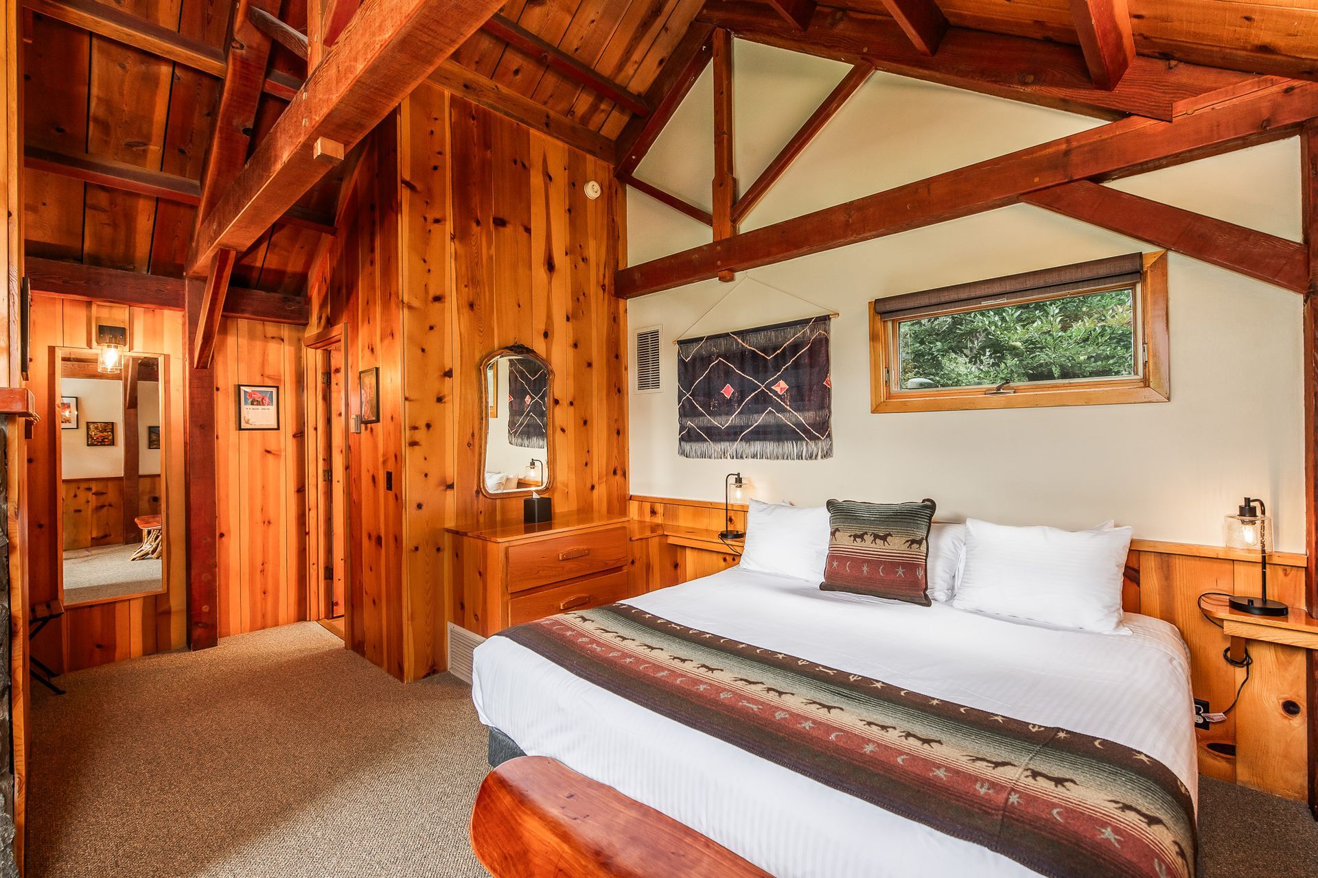 Cozy bedroom with wooden walls and ceiling, a bed with a patterned blanket, and a view from the window.
