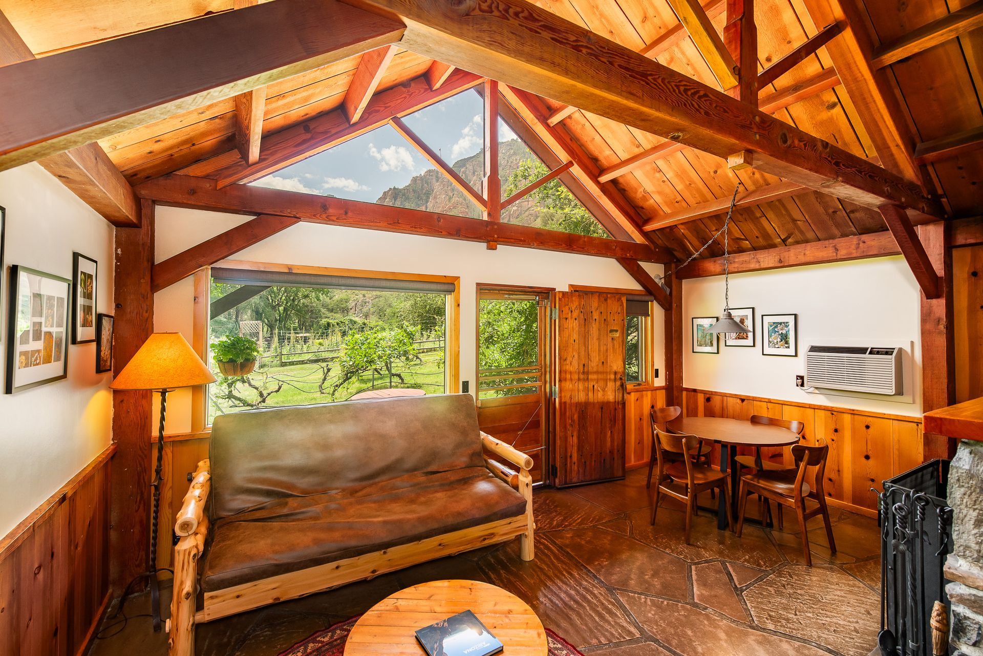 Cozy cabin interior with wooden beams, futon, dining table, and a view of lush greenery outside the windows.