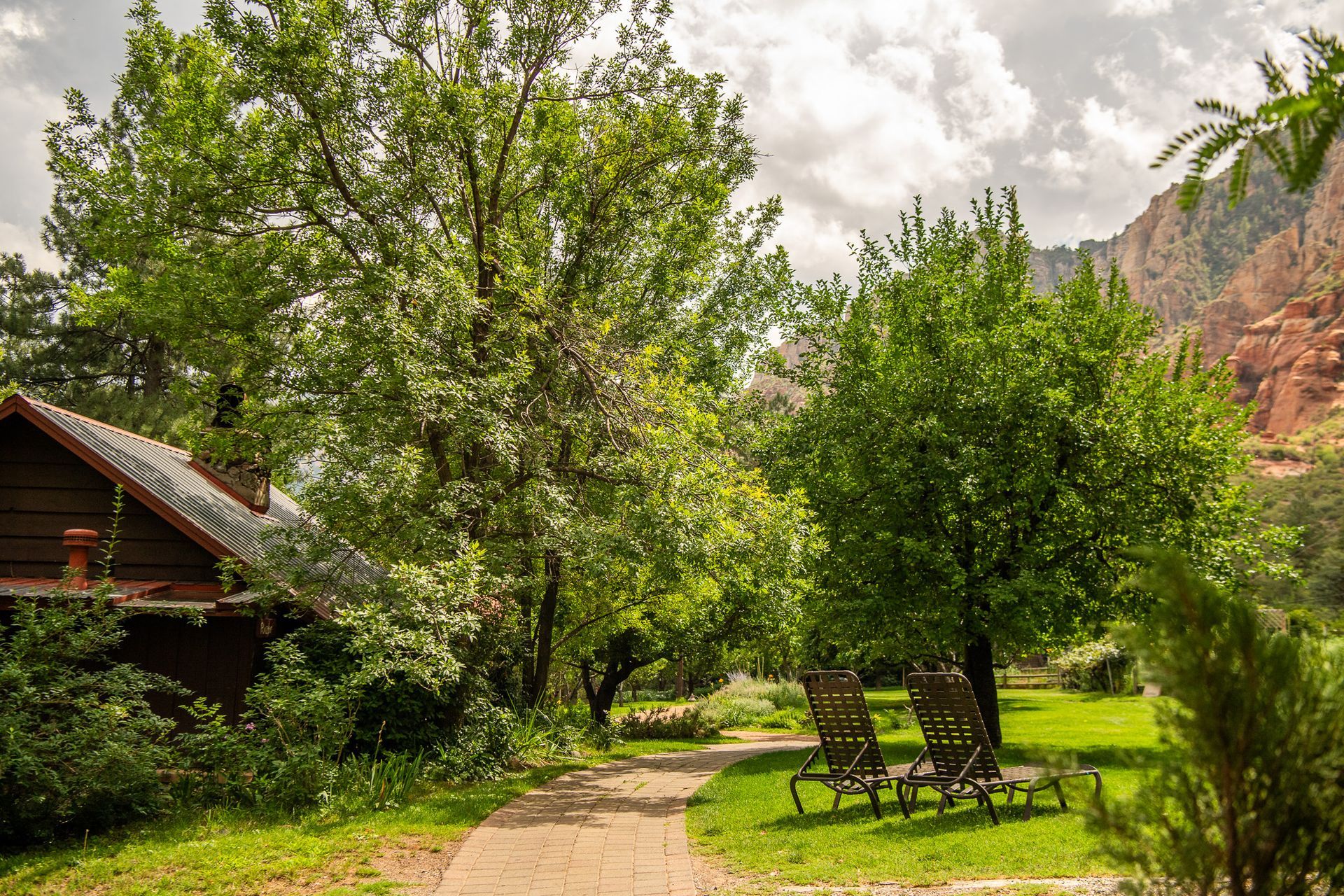 Stone path leads past trees to two chairs on a lawn; a house and red rock canyon in the background.
