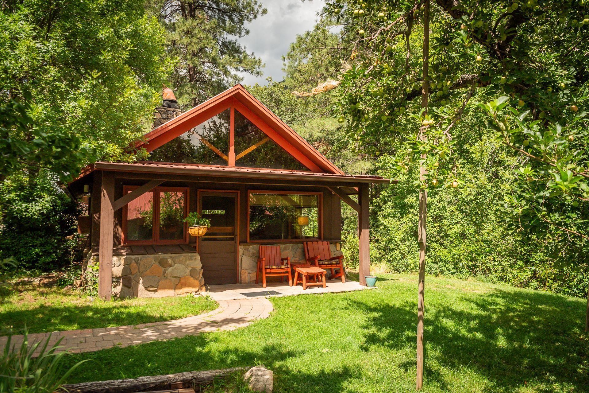 Cozy cabin with stone base, porch, and red roof, surrounded by trees and lawn.