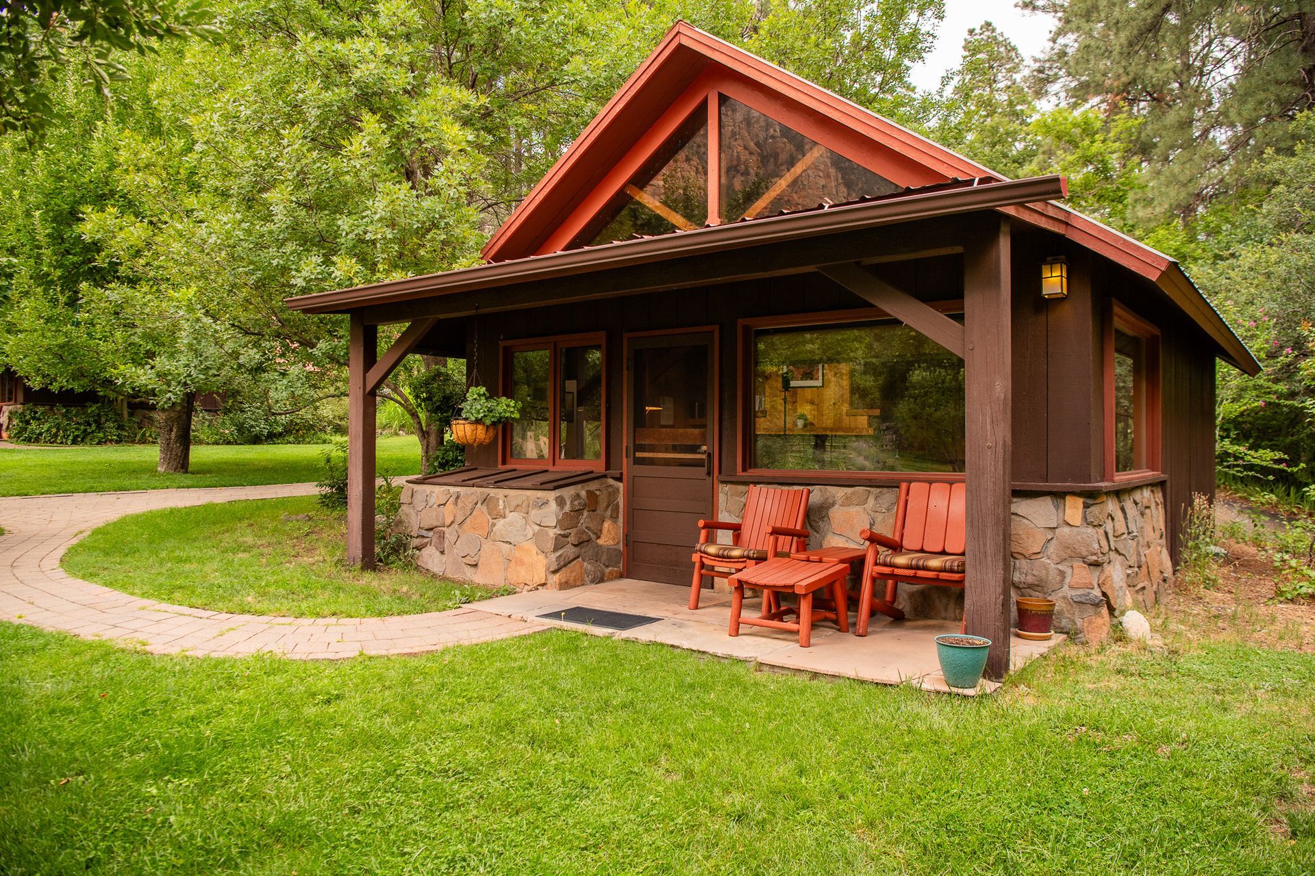 Small brown cabin with stone base, porch, and red chairs on a green lawn with a brick pathway.