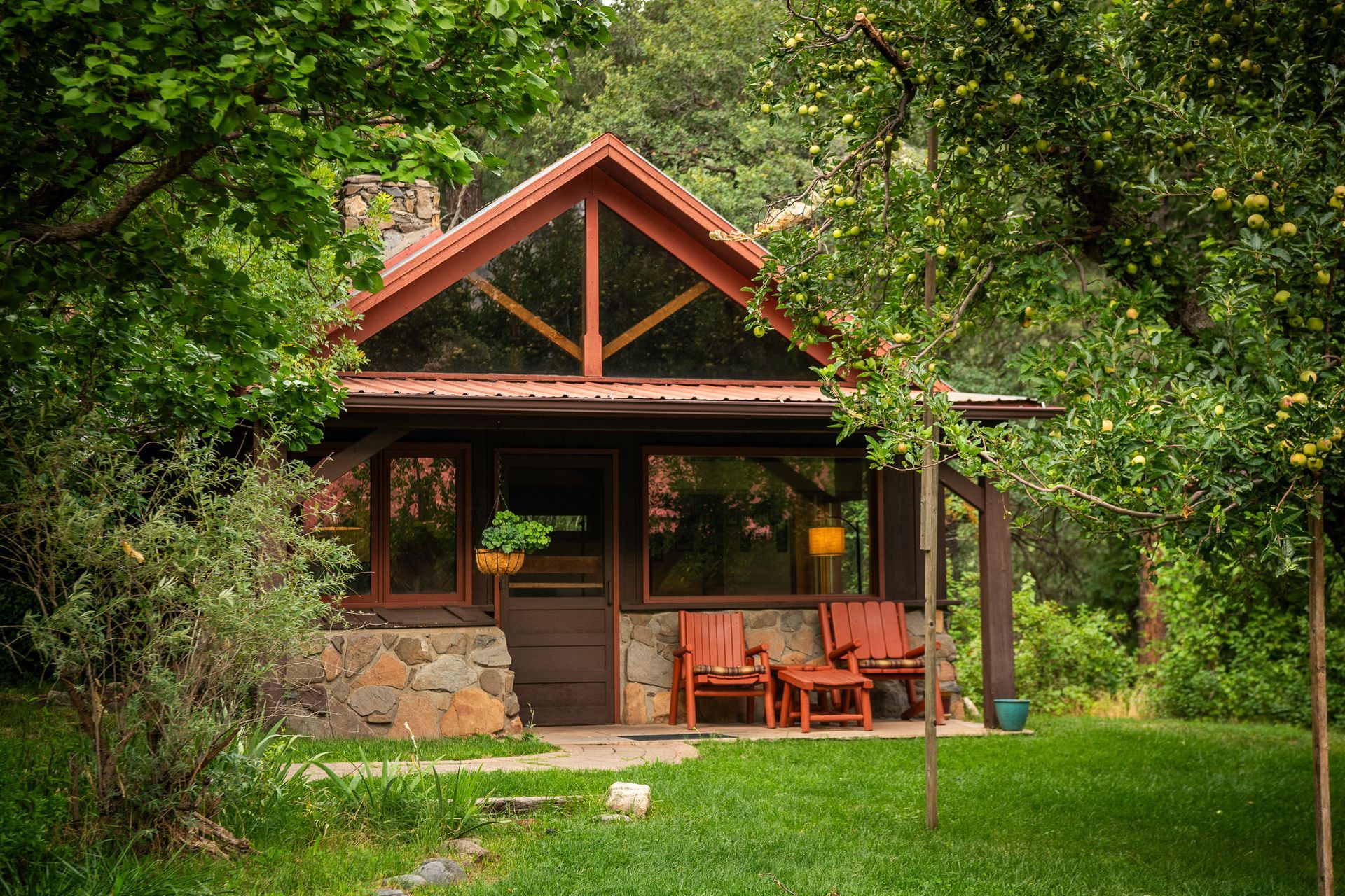 Cozy cabin nestled in a lush green yard, with a stone base, a porch, and two orange chairs.